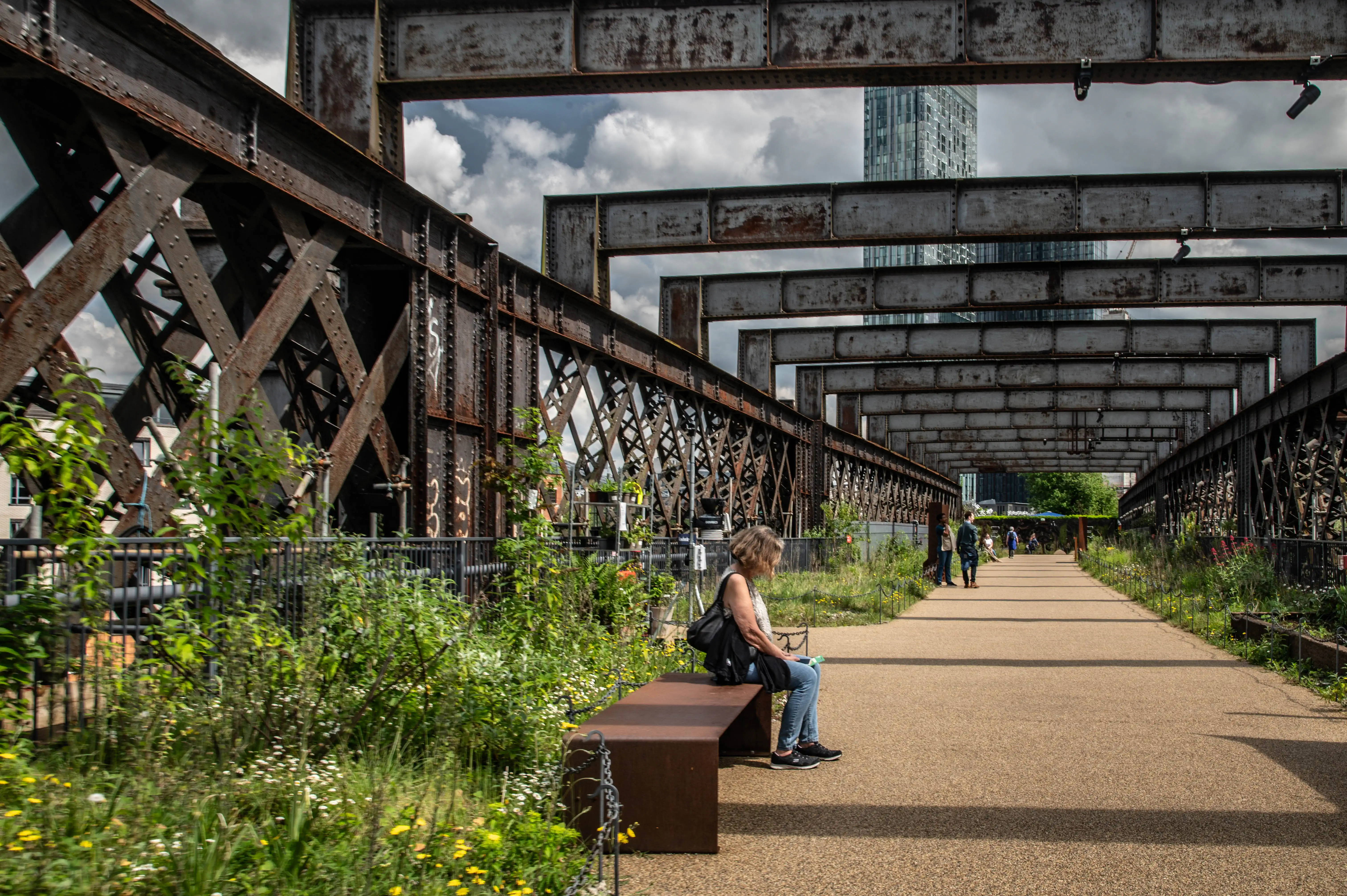  A woman is seated on a rustic metal bench on a wide, sandy-coloured pathway. The pathway is flanked by lush green plants and flowers on one side and the imposing, rusty steel structure of an old railway bridge on the other. In the distance, more of the elevated structure recedes, leading towards a modern, glass-fronted skyscraper under a cloudy sky.