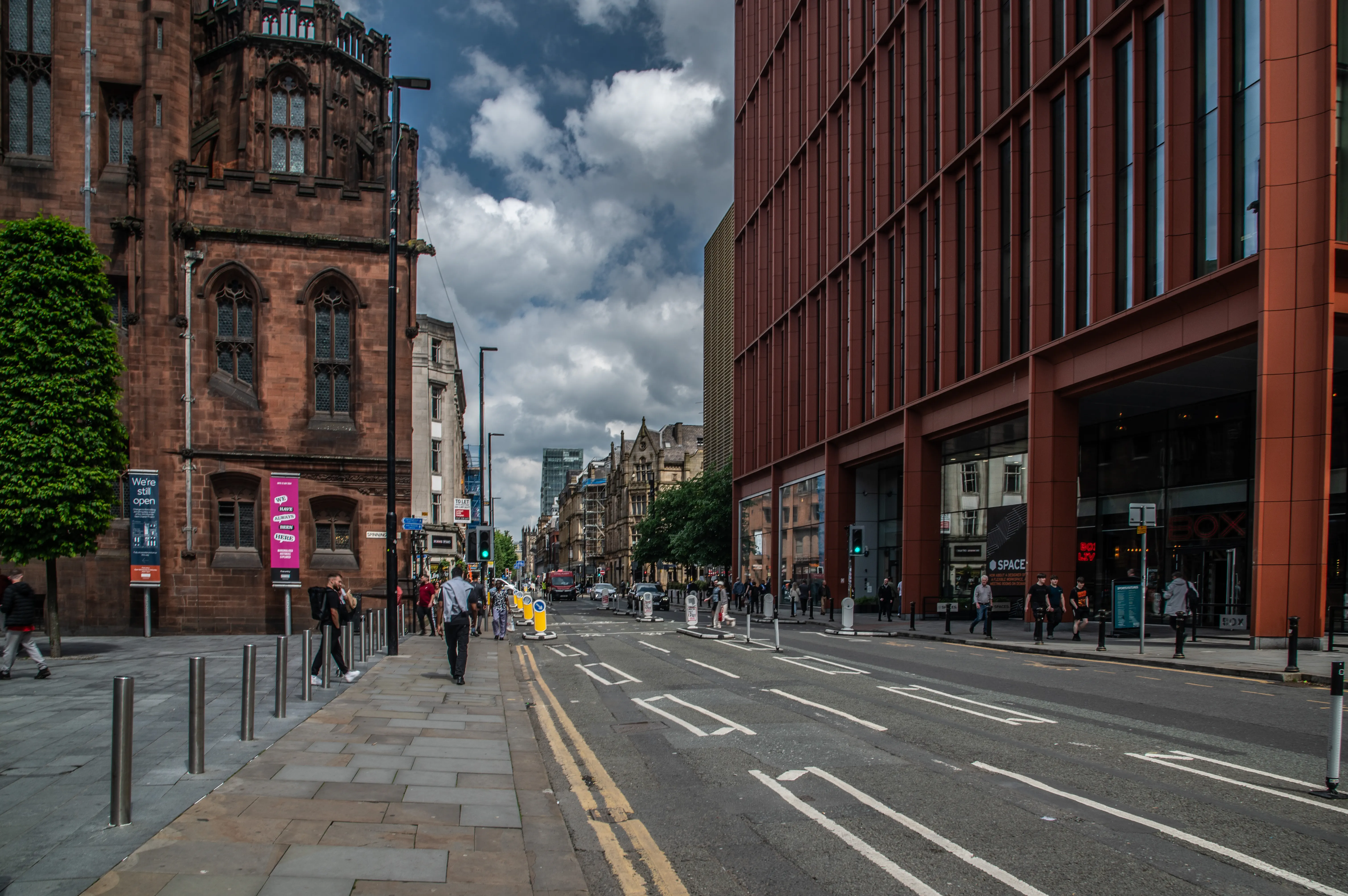 A wide street with a modern red-brick building on the right and an older stone building on the left, with people walking on the pavement and cars on the road under a cloudy sky.