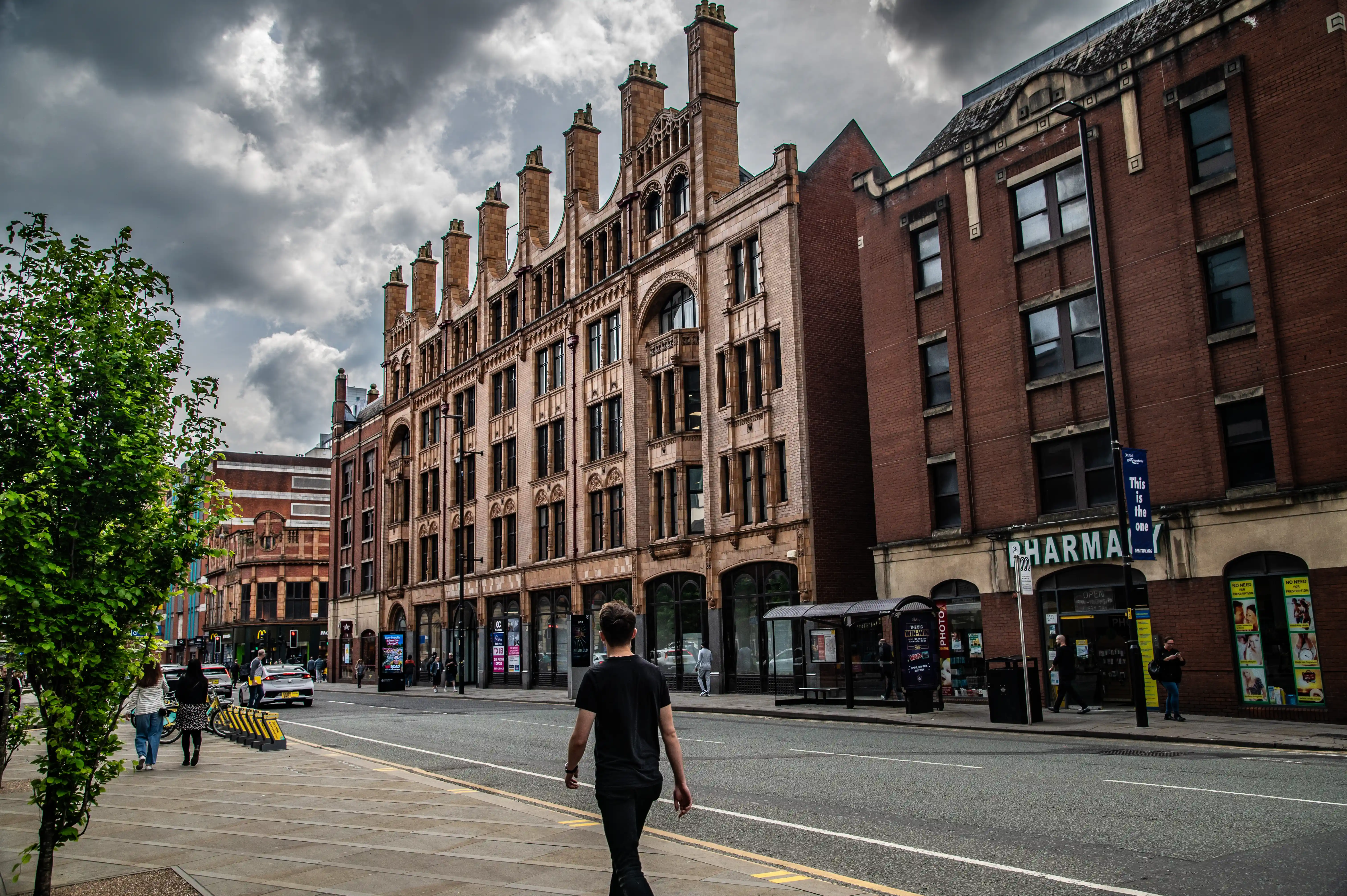 A young man walks away from the camera on a city pavement, with ornate brick buildings and a pharmacy lining the street under a dark, cloudy sky.