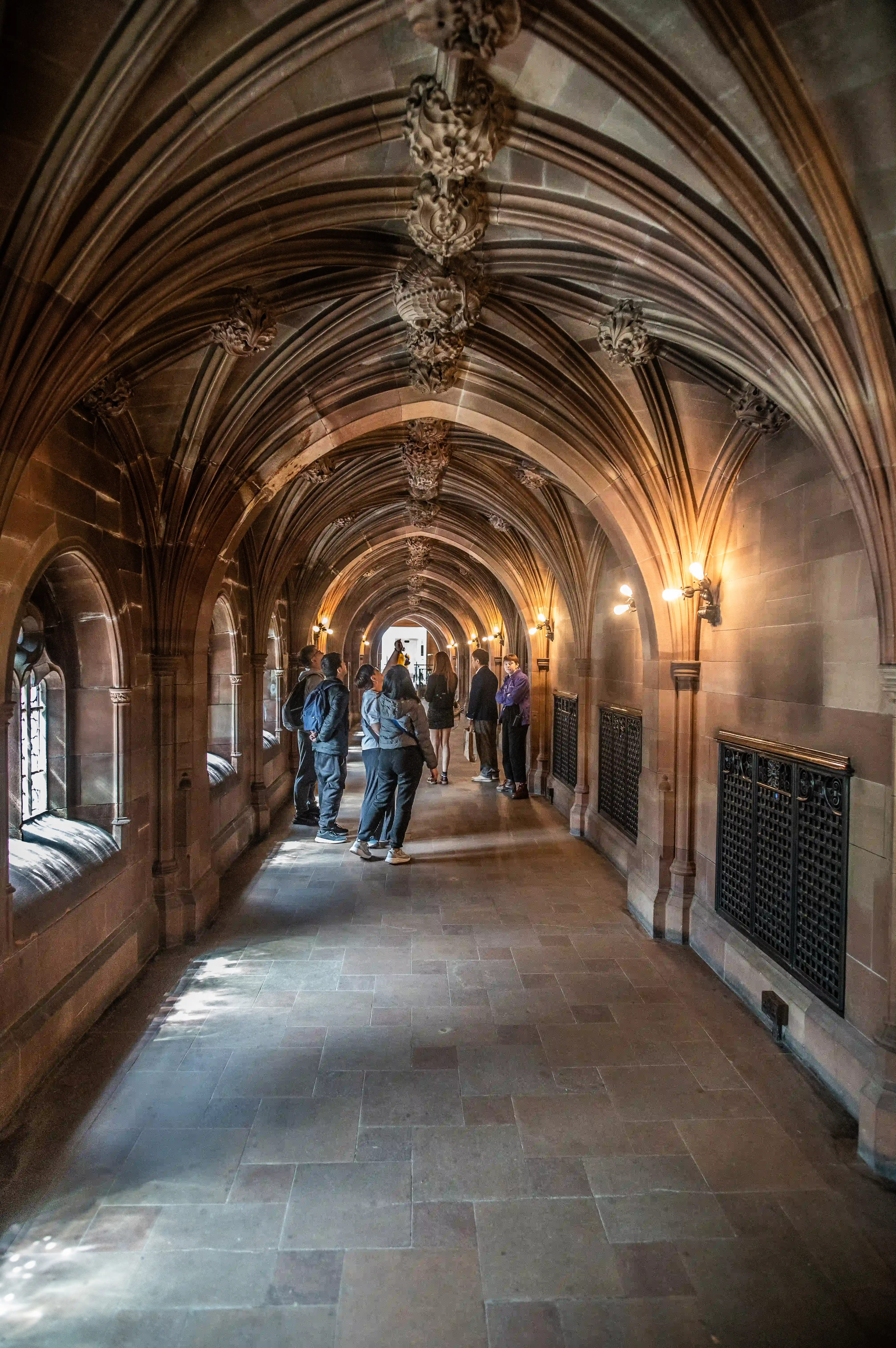 A perspective view of a long, stone hallway with a high, vaulted, and intricately decorated Gothic ceiling. Several people are walking down the hallway, and light streams in from arched windows on the left.