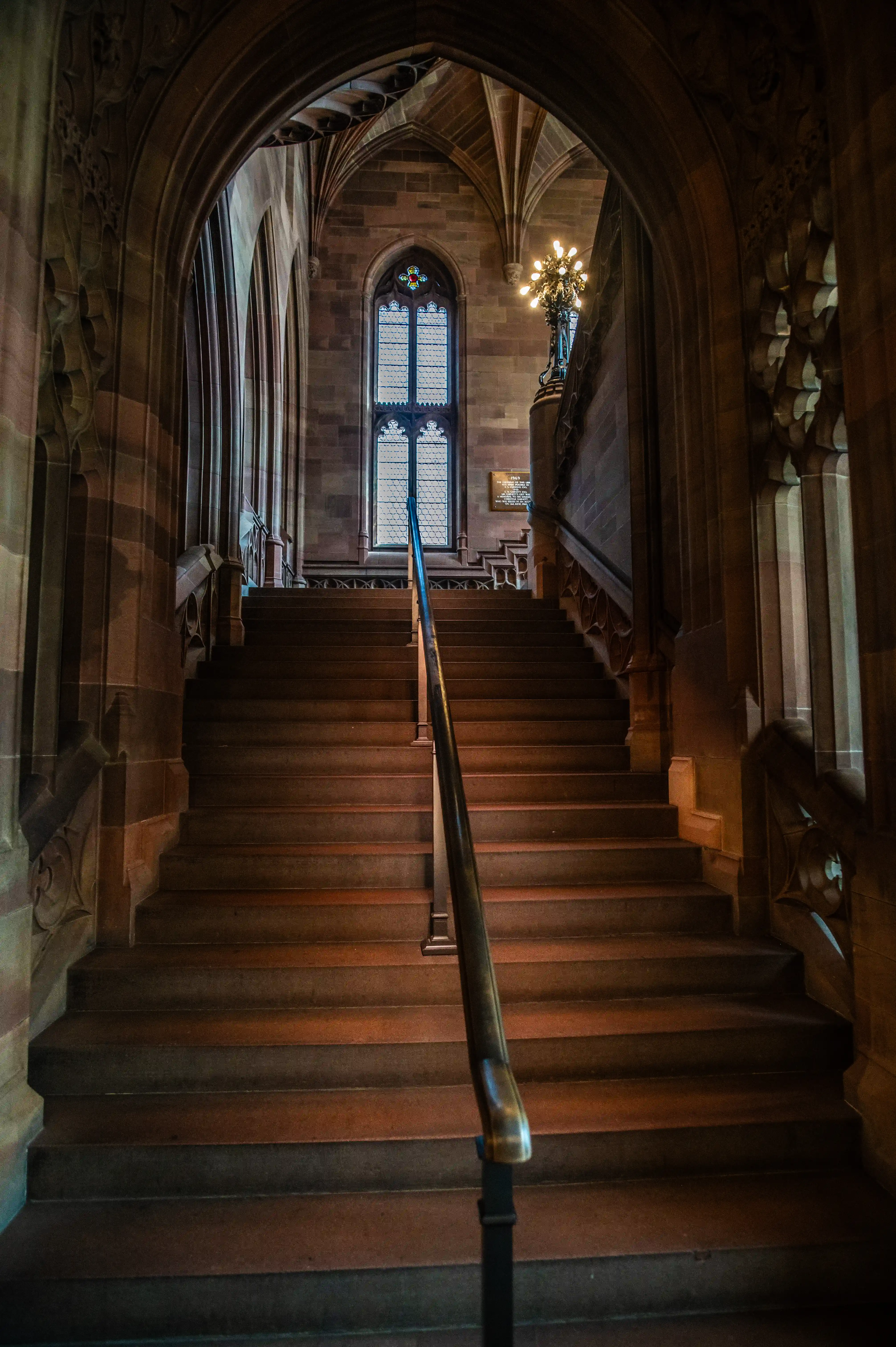 A wide, symmetrical view of a grand stone staircase with a dark wooden handrail leading upwards. The staircase is framed by intricate stone arches and columns with Gothic detailing. At the top of the stairs, a large leaded window is visible, and a decorative chandelier hangs nearby