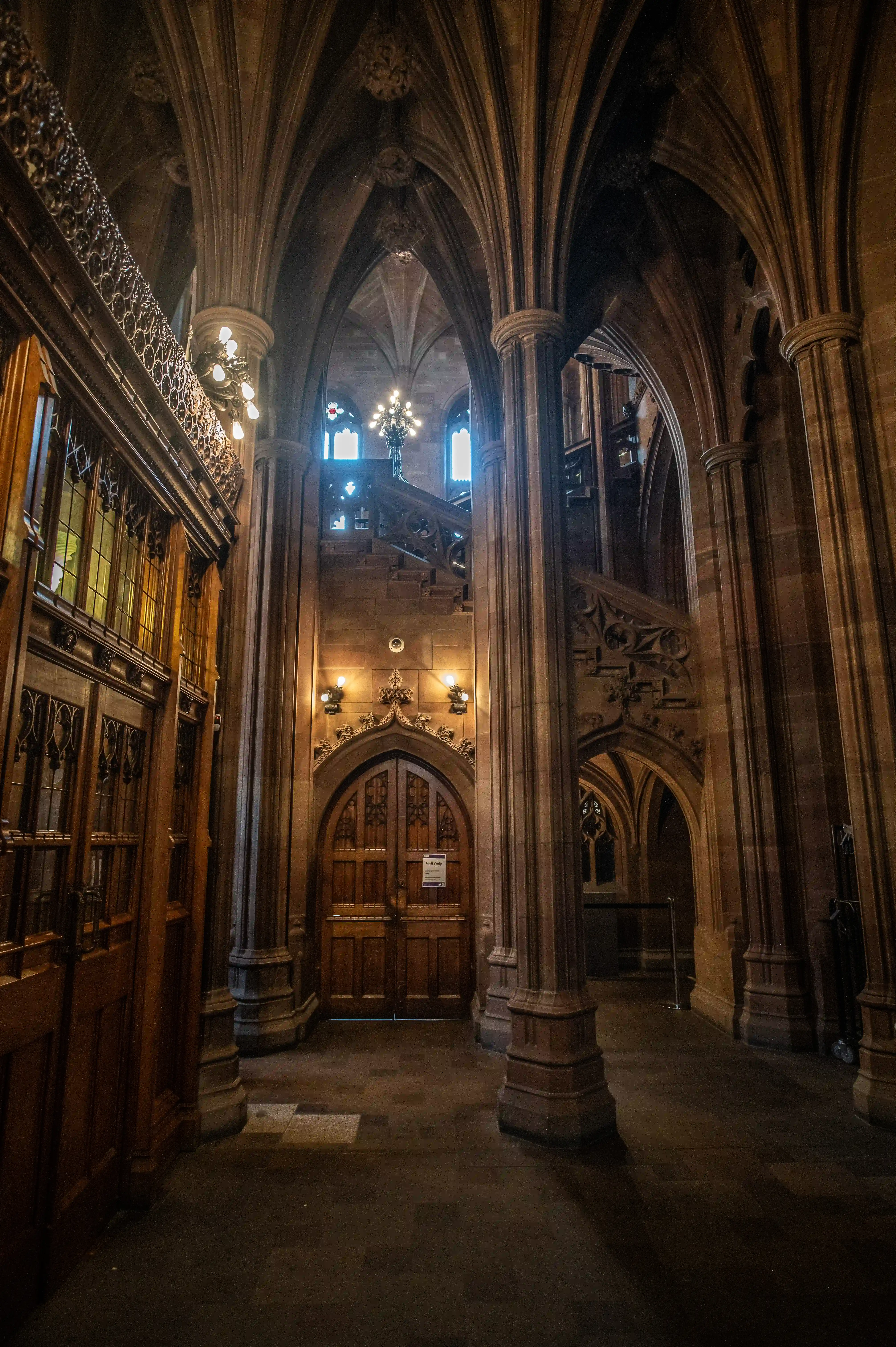 Gothic interior with pointed arches, ribbed vaults, carved stone columns, stained-glass door, and a stone staircase.