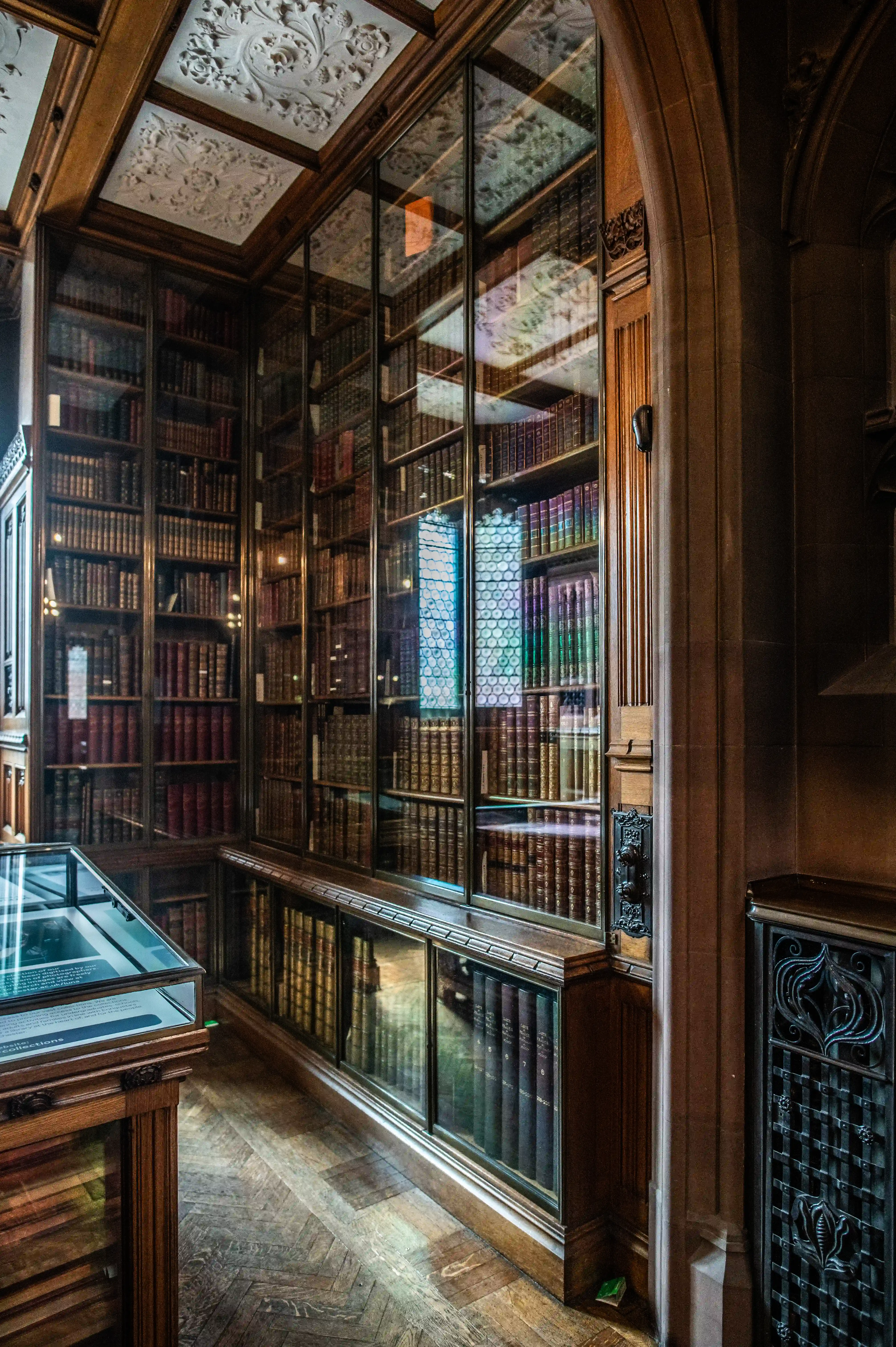 Ornate library with tall glass-fronted wooden bookcases, leather-bound books, plaster ceiling, and a glass display case.