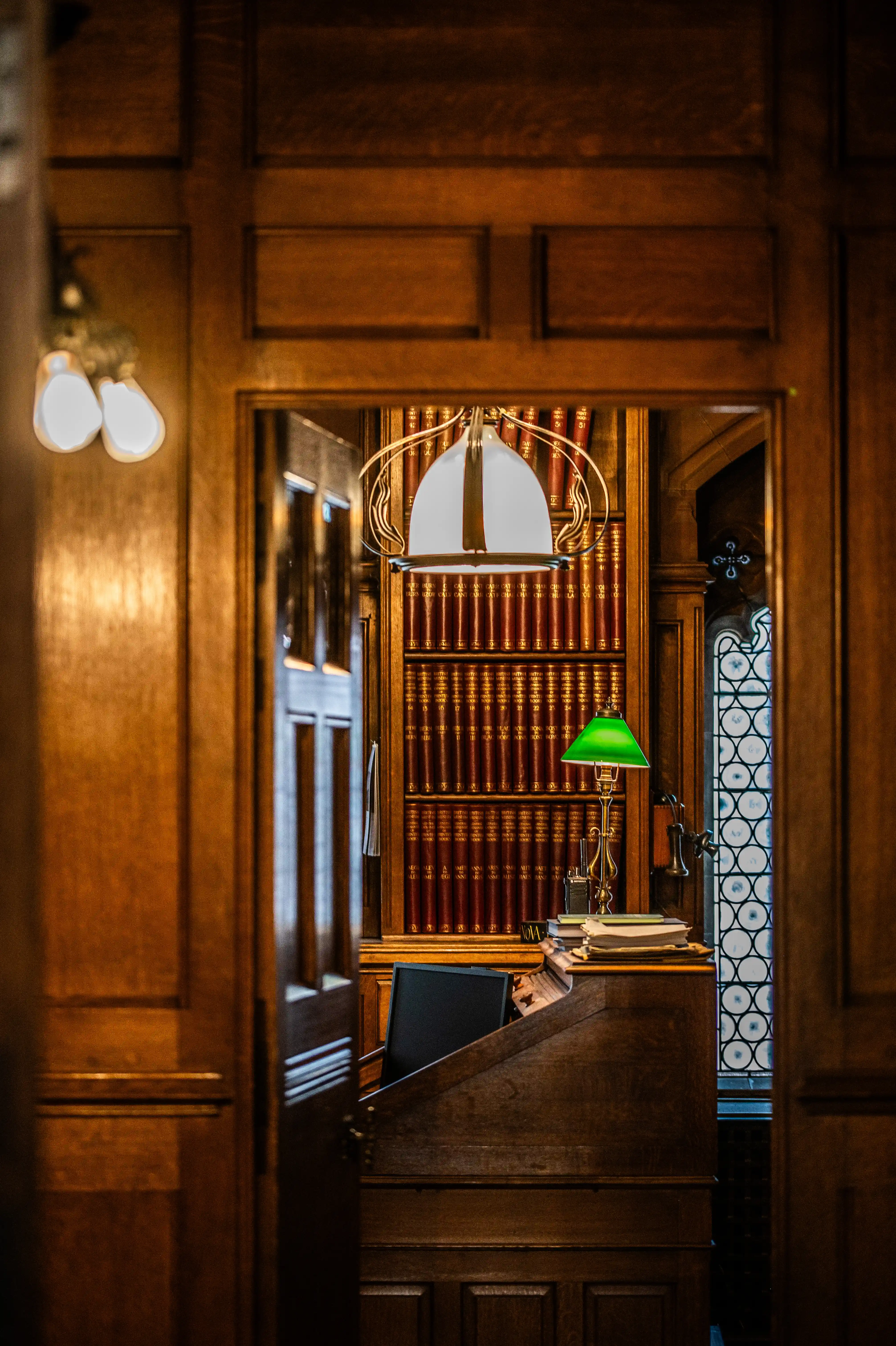 Historic library interior showing a writing desk with lamps and stacked books, framed by wood panelling and tall shelves