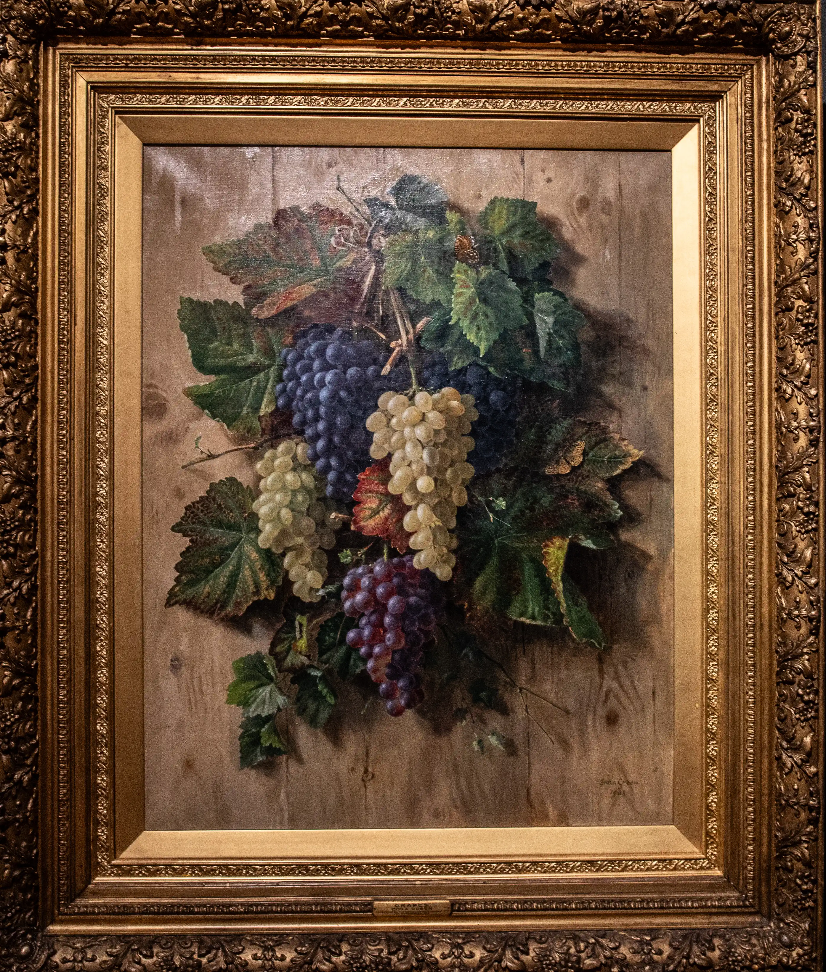 Framed still life painting of purple and pale grapes with green vine leaves on a wooden-textured backdrop.  