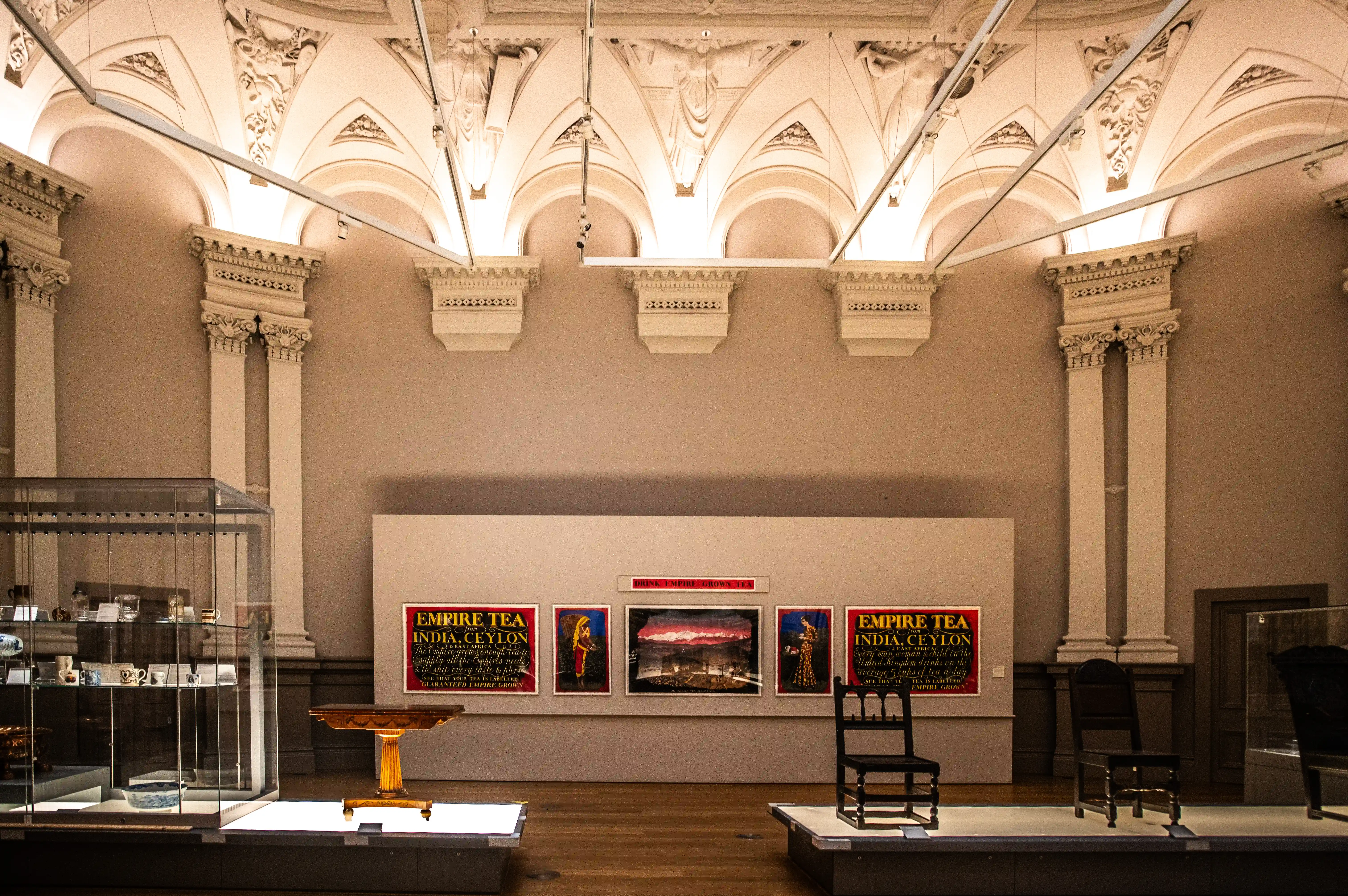Interior view of a museum exhibition hall, displaying vintage posters for 'Empire Tea' from India and Ceylon, a glass display cabinet filled with porcelain, a small ornate table, and antique chairs.