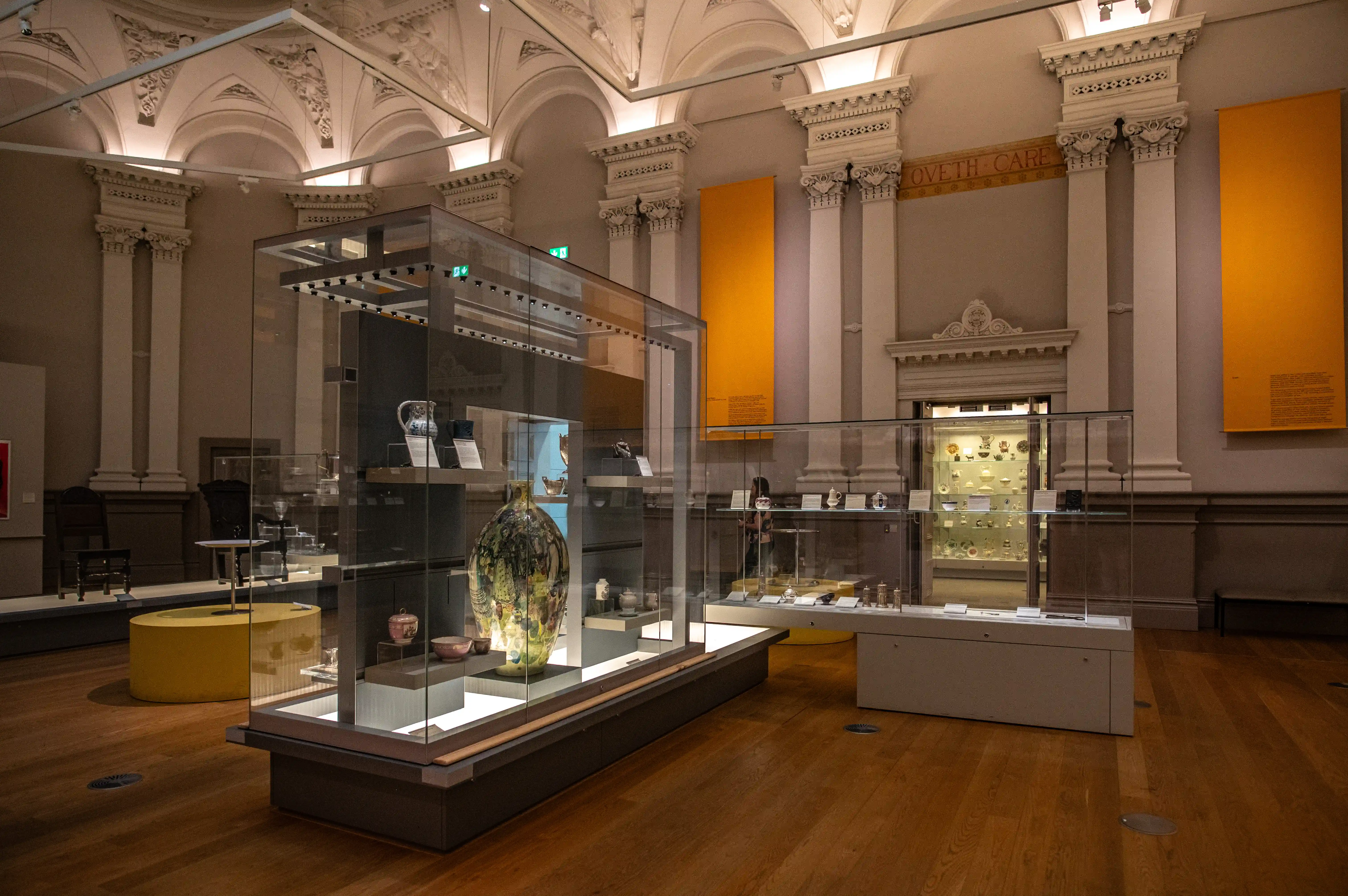 A museum gallery with glass display cases containing historical artifacts like pottery, vases, and decorative objects. The room has tall columns, ornate moldings, and yellow informational panels, with soft lighting highlighting the items.