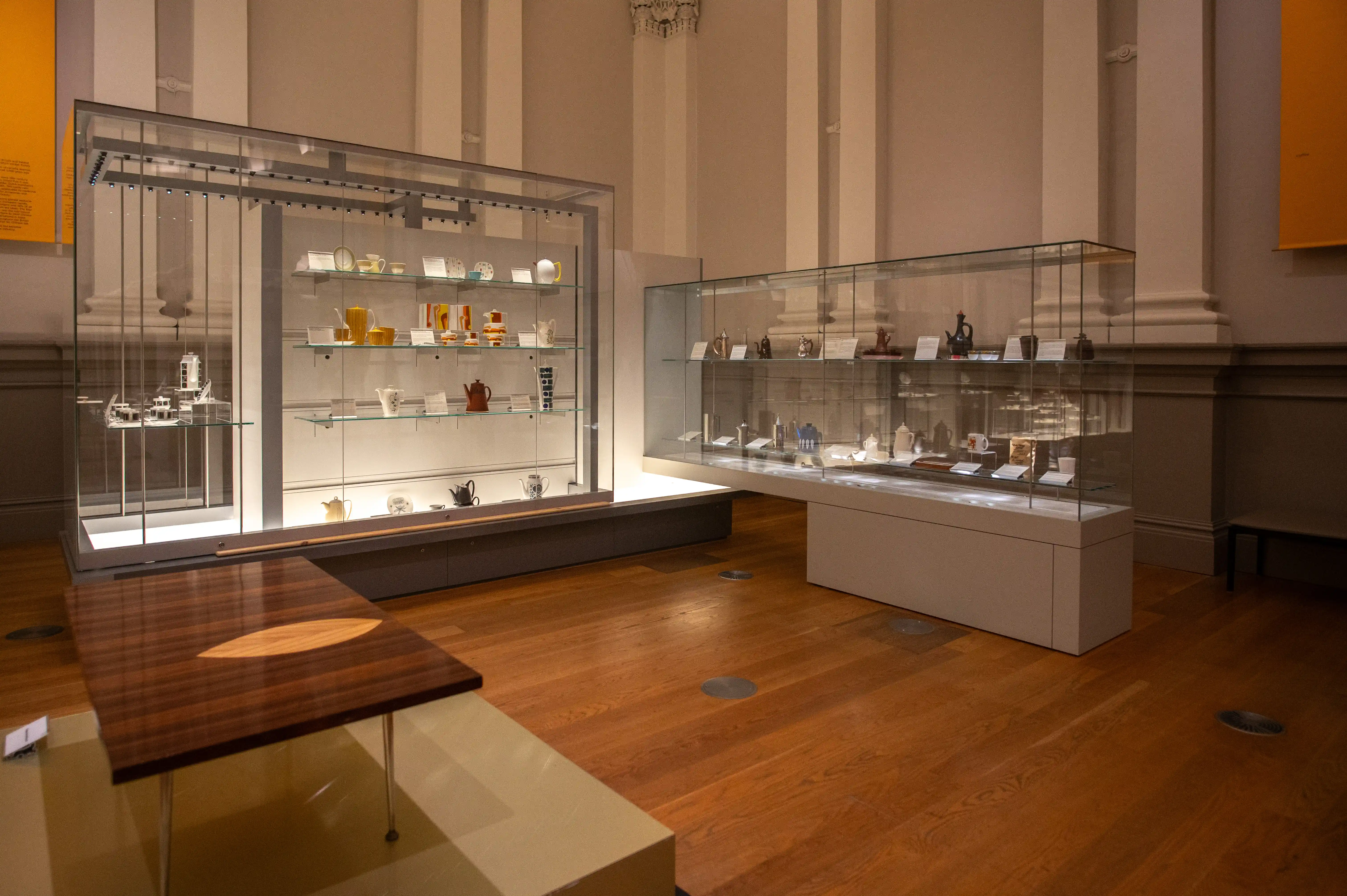 A museum gallery with glass display cases containing historical artifacts like pottery, vases, and decorative objects. The room has tall columns, ornate moldings, and yellow informational panels, with soft lighting highlighting the items