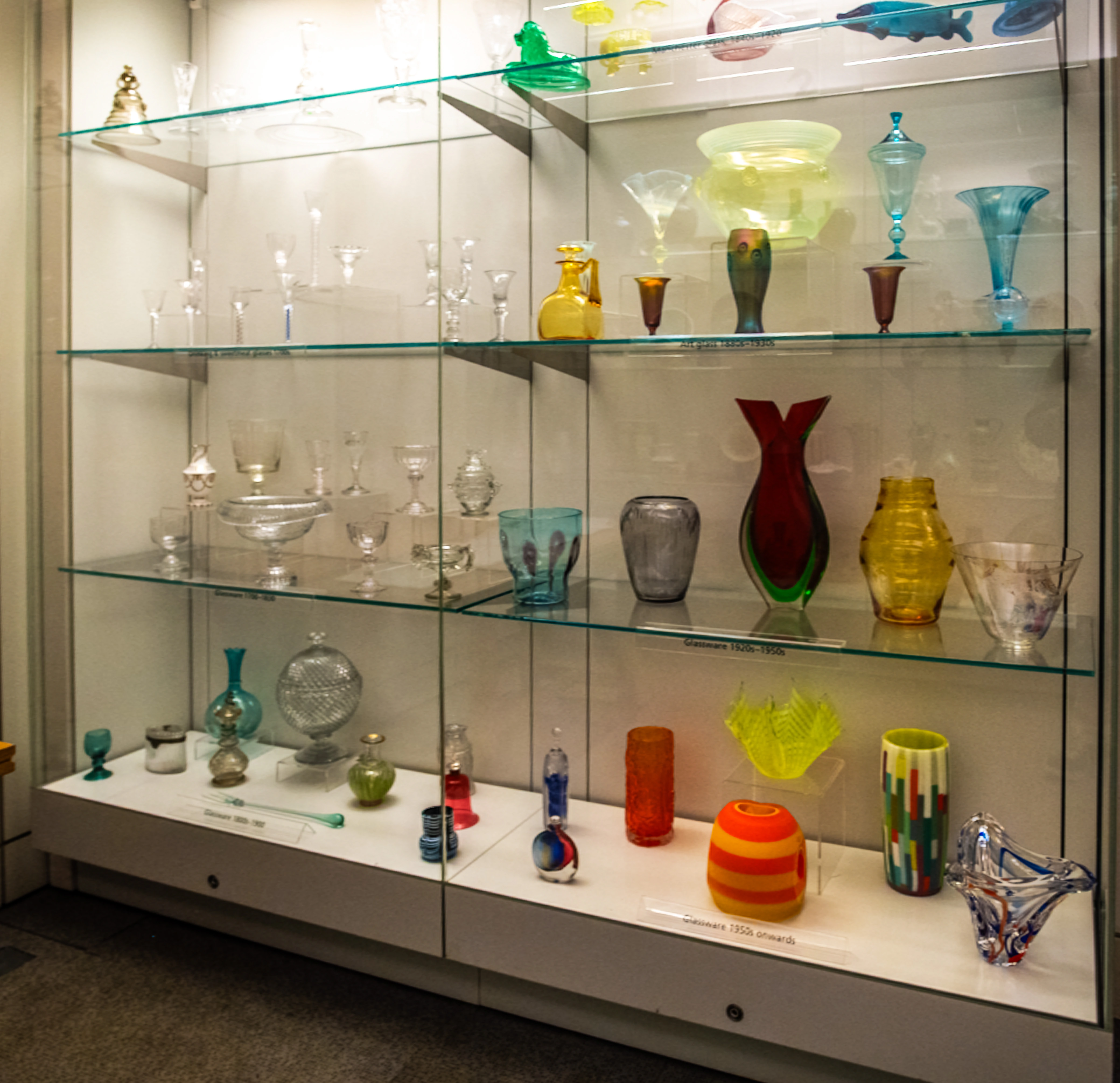 Backlit museum display case with multi-coloured glass vessels, vases, and goblets arranged across shelves. 