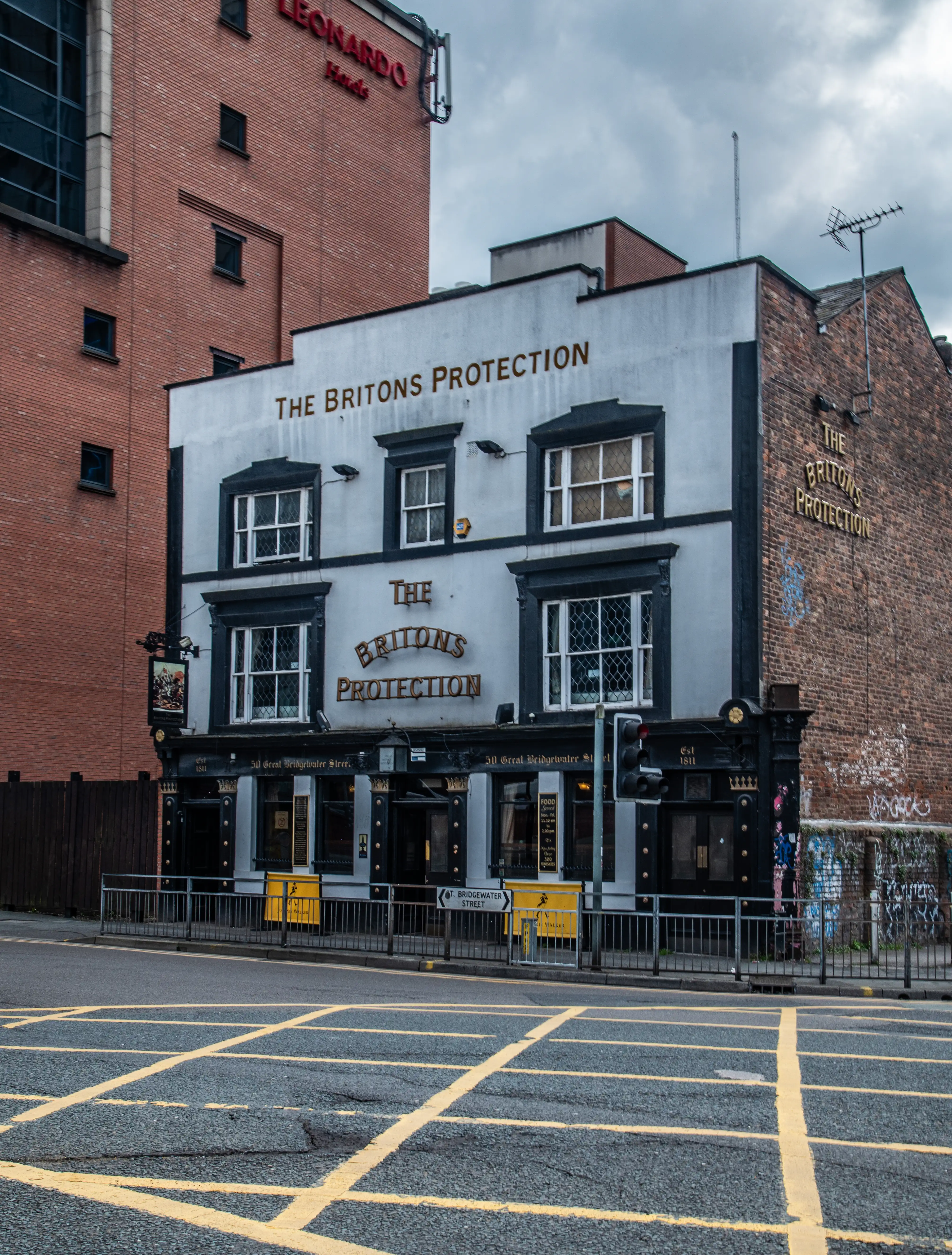 Front of a building painted with the words “THE BRITONS PROTECTION”, with windowed bays and a surrounding brick wall on a street.