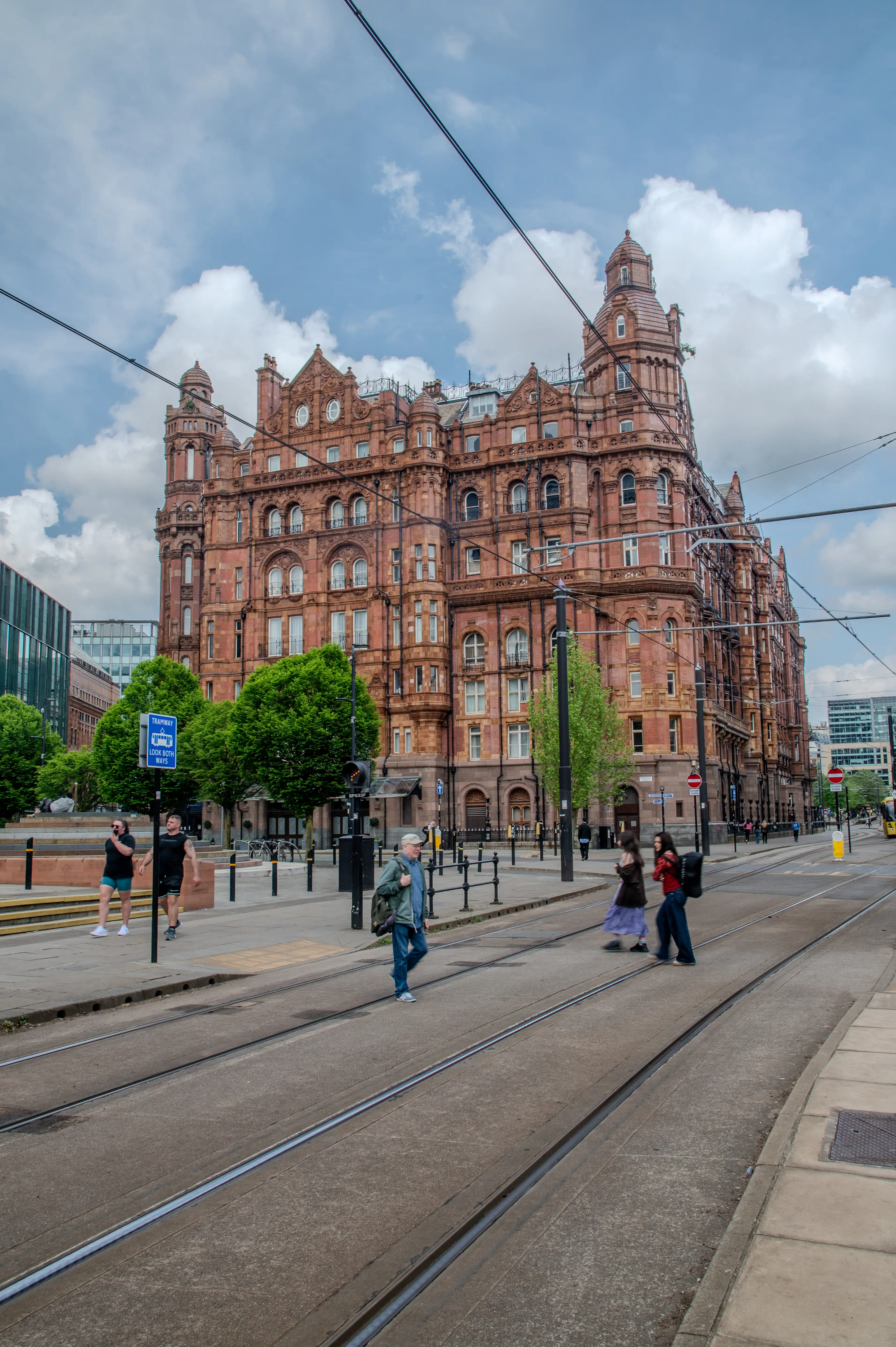 Red-brick historic building with towers, viewed from street level beside tram tracks, with pedestrians walking under a cloudy sky. 