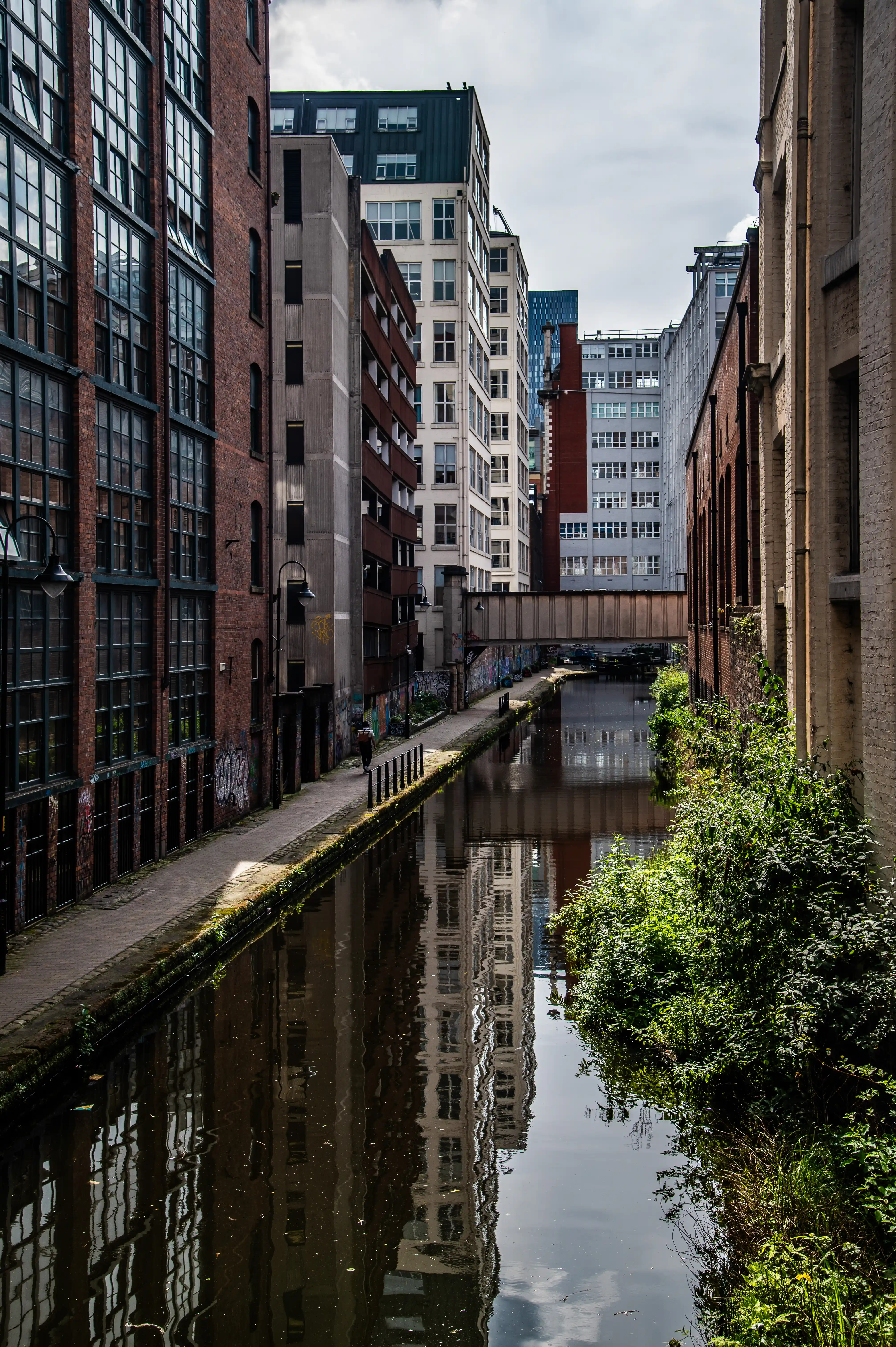 View down a narrow canal between tall brick buildings and modern offices, with reflections on the water and cloudy sky above.  