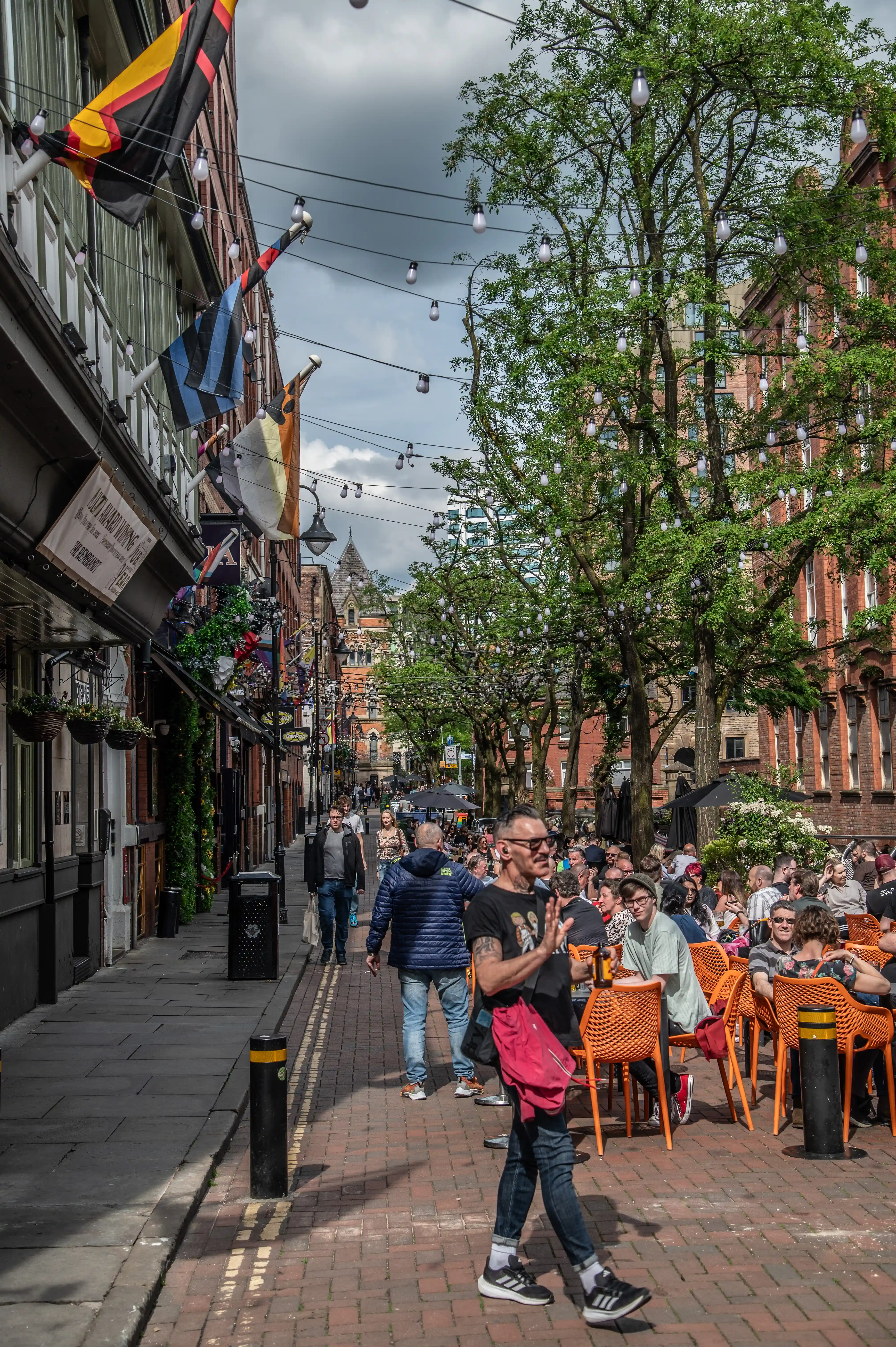 Outdoor seated crowd on a narrow brick street with orange chairs, hanging bunting flags, and string lights above trees.