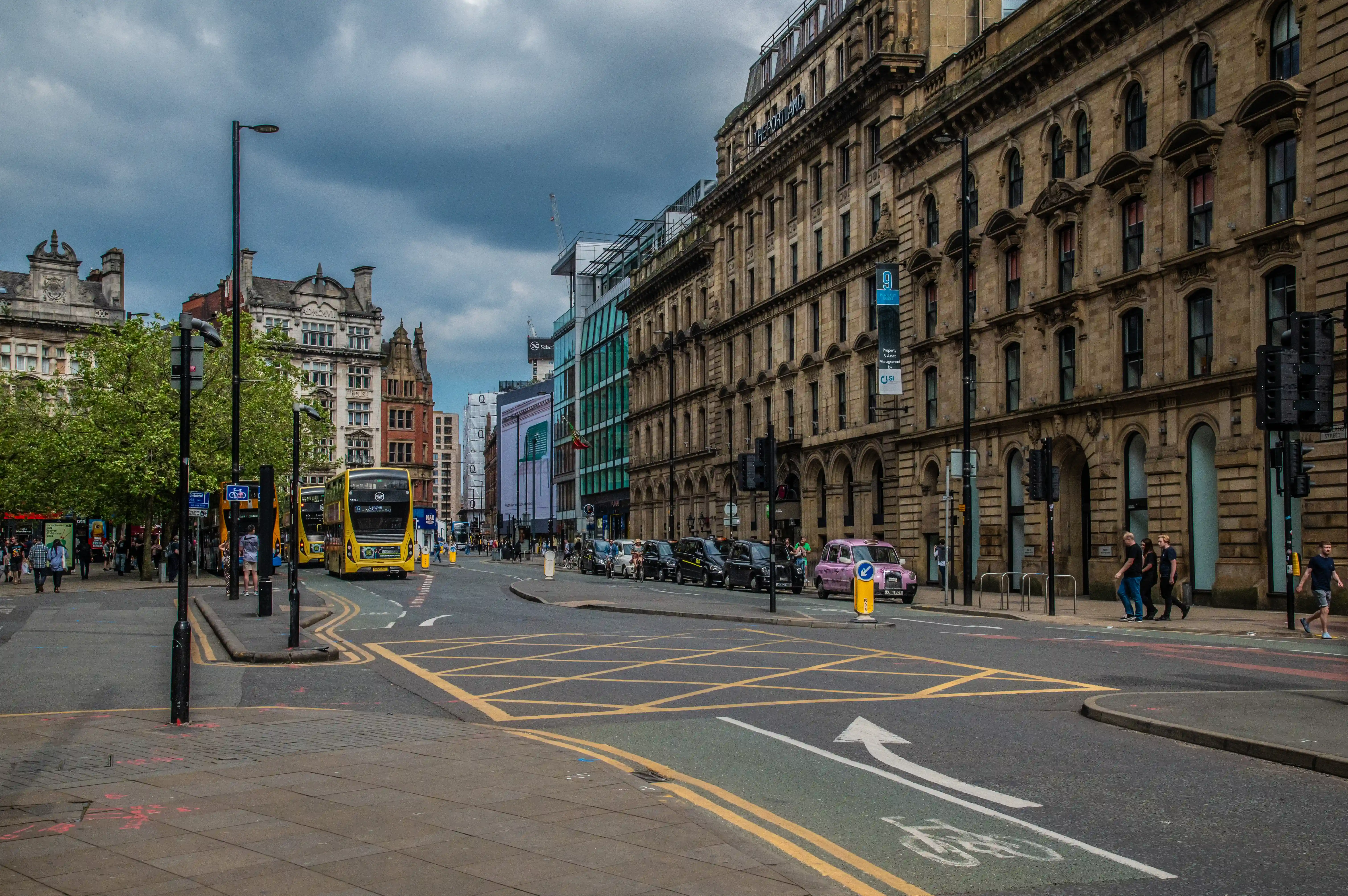 A street in Manchester city centre with yellow double-decker buses, cars, pedestrians, and buildings of varying architectural styles.