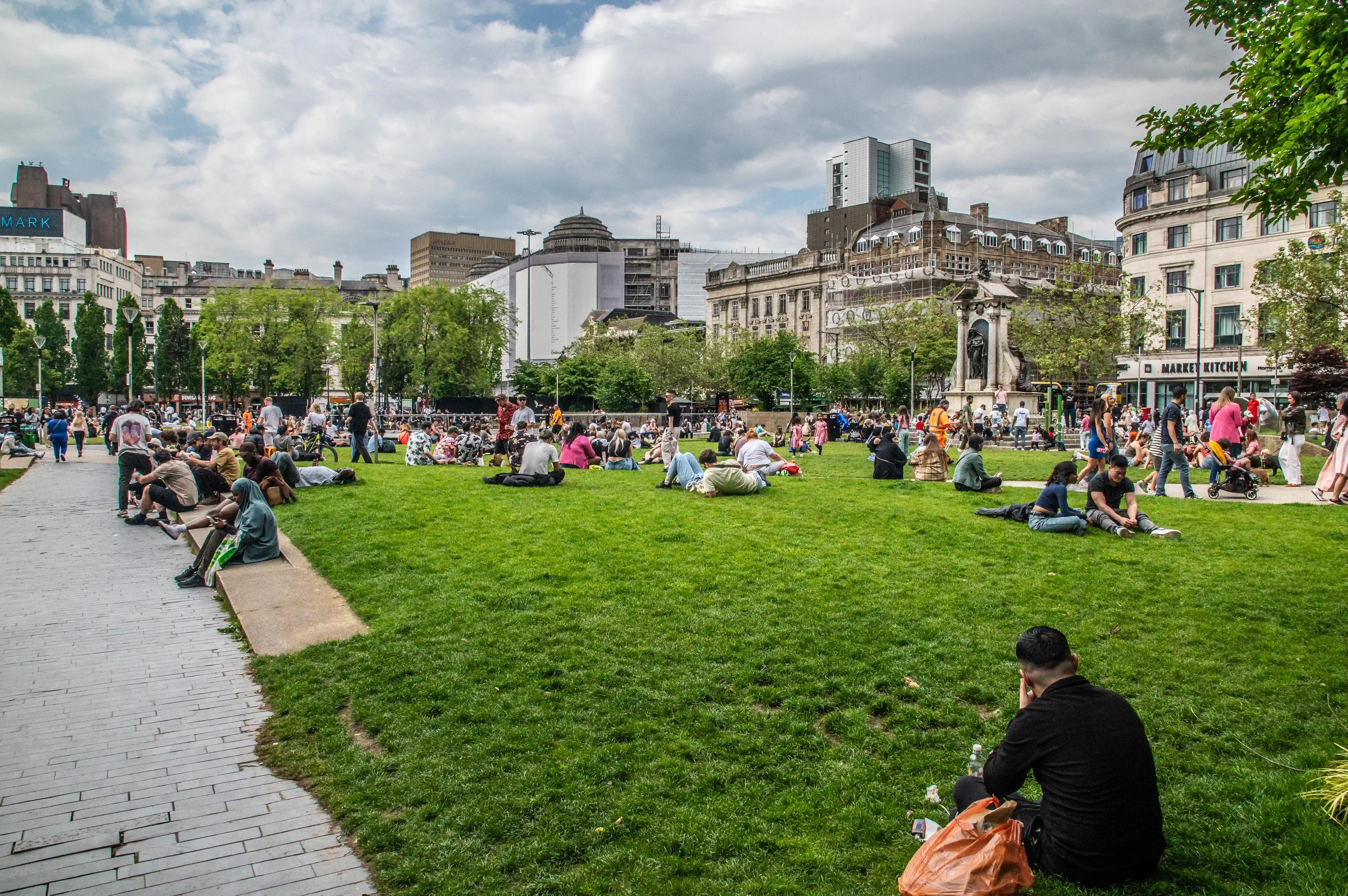 People sitting and lounging on a large grassy area in a city park, with buildings visible in the background.