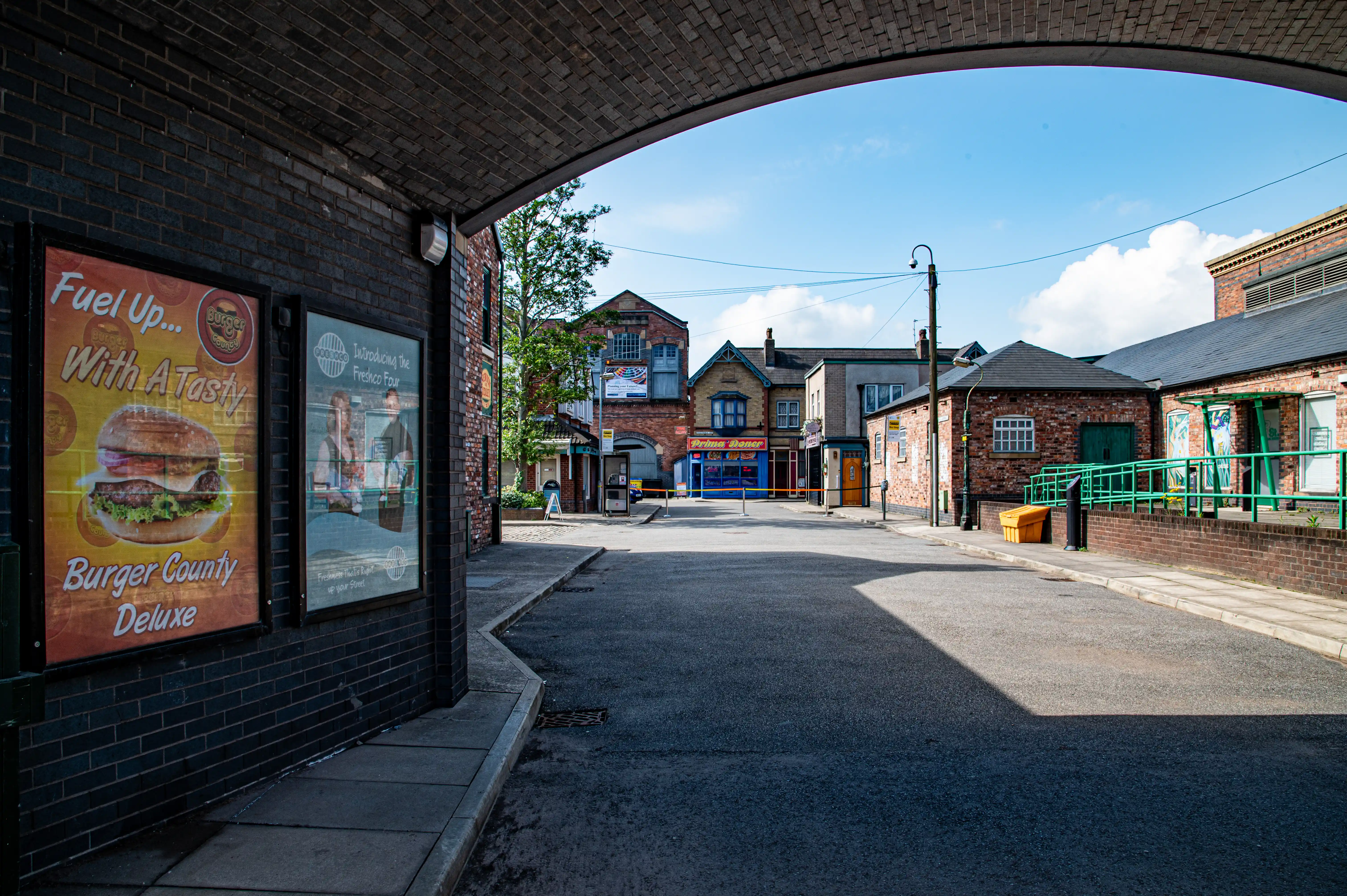 A street scene from the Coronation Street television set, with brick buildings lining both sides and an asphalt road. A prominent archway frames the view of the street which recedes into the distance. Two large posters are displayed on the left wall, one advertising 'Burger County Deluxe'.