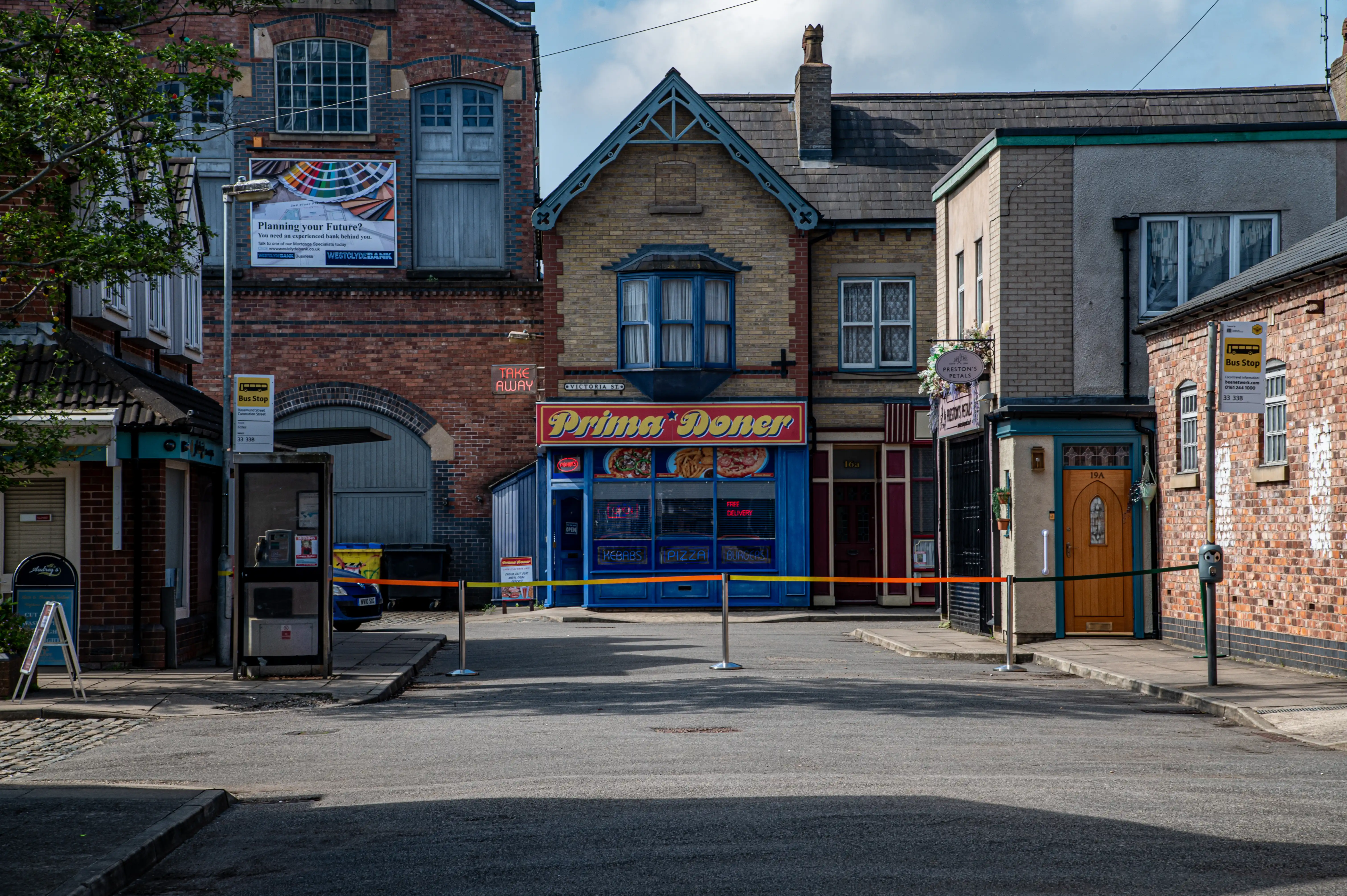 A view of a street lined with traditional brick buildings housing various businesses, including a doner kebab shop and a bus stop, with a road in the foreground.