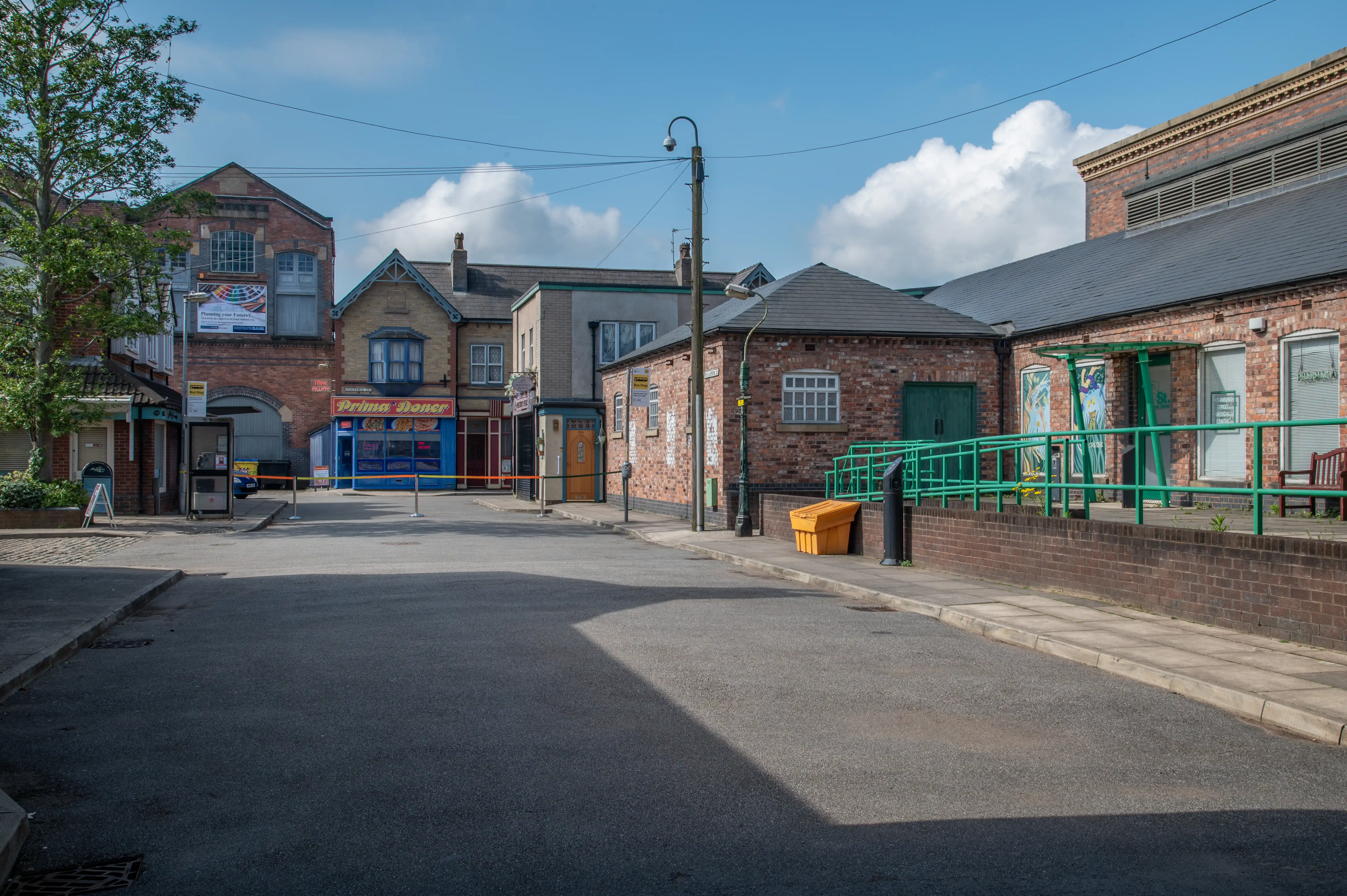 A wide shot of a British street scene, featuring several brick buildings with shopfronts, a paved road, and a lamppost. The sky is blue with white clouds.