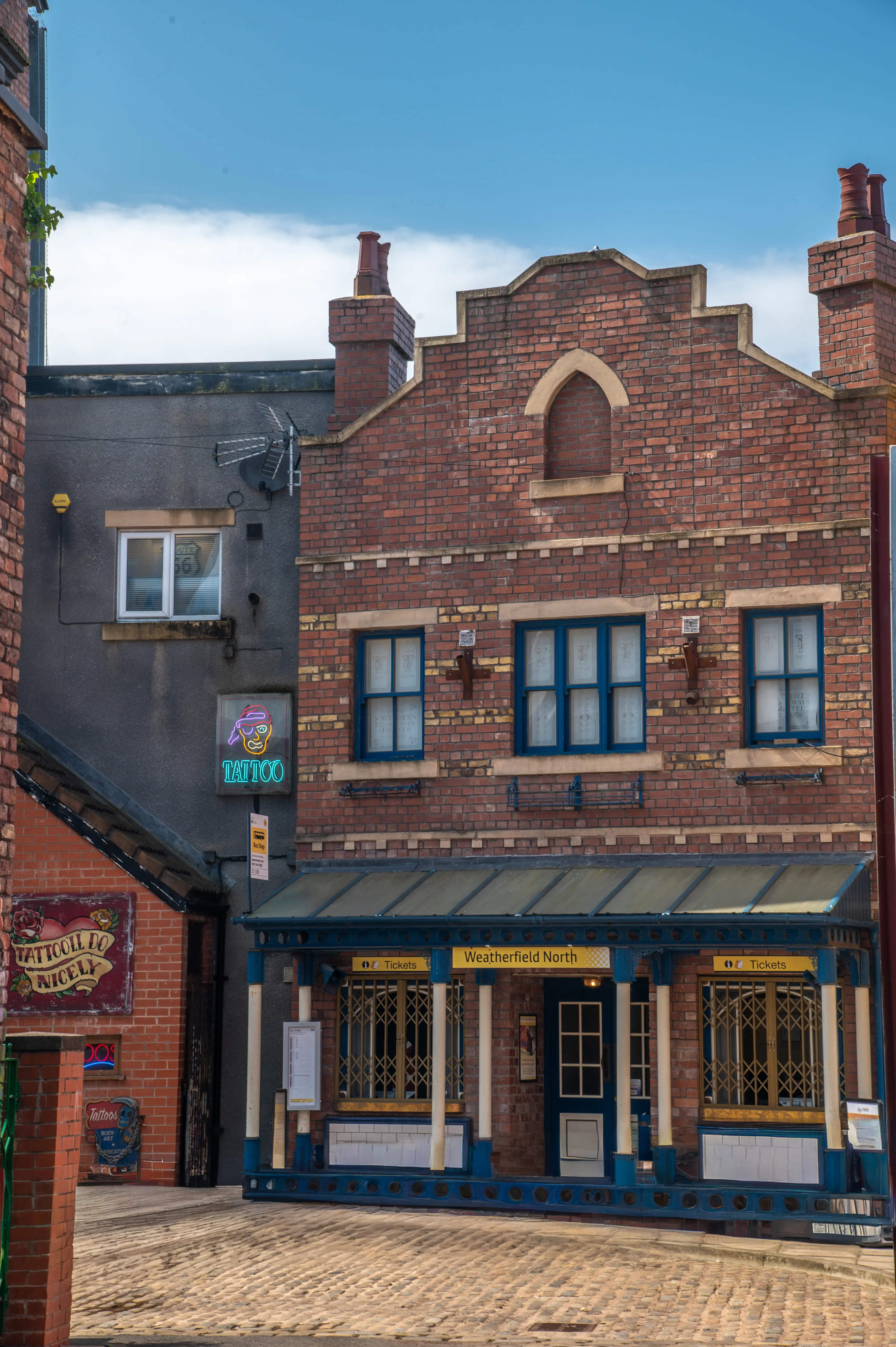 Exterior view of Weatherfield North train station with blue and gold trim and ticket windows, next to a grey building with a glowing neon tattoo sign, under a blue sky with scattered clouds.