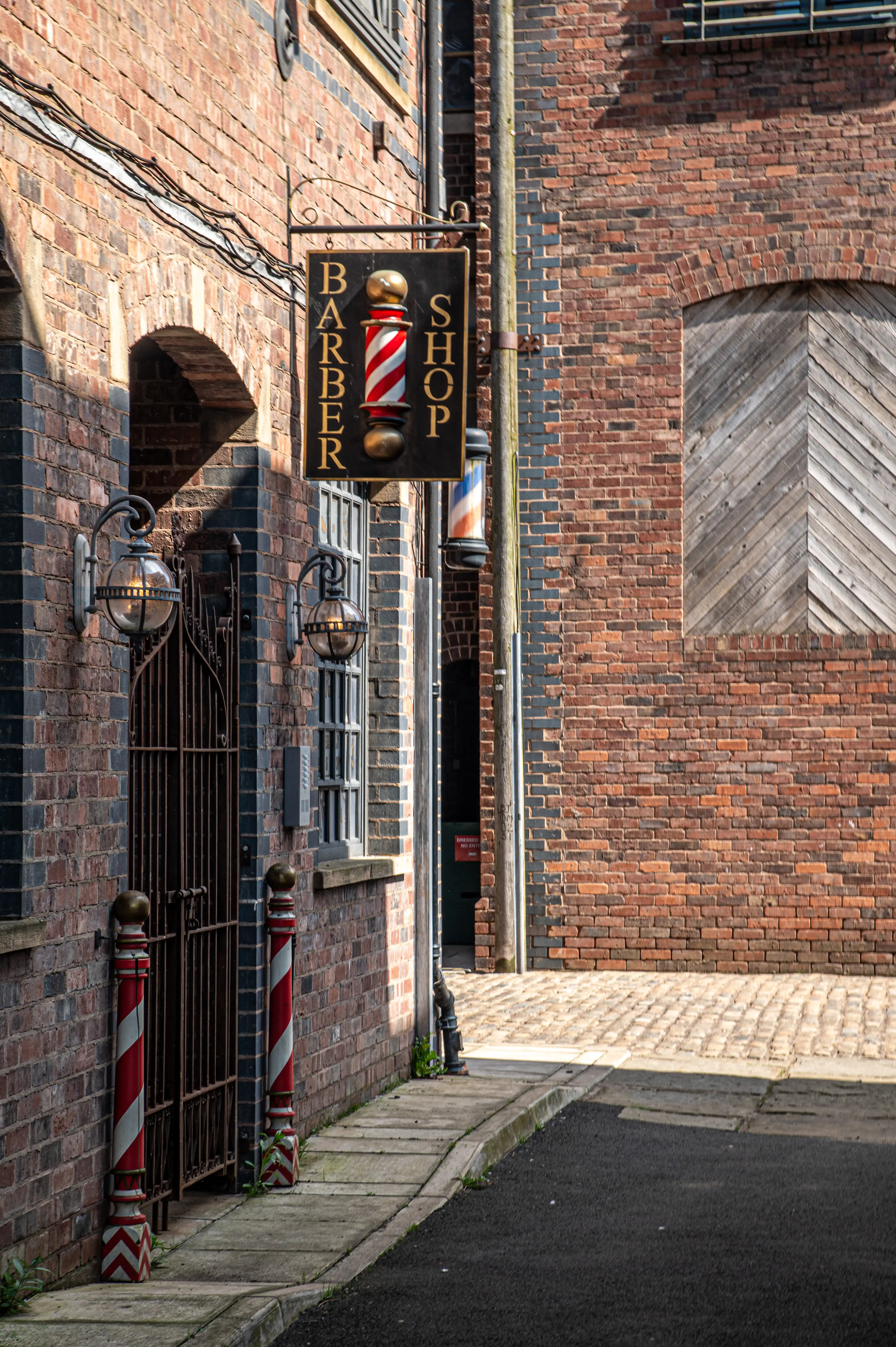 A vertical barber pole sign reading &laquo;BARBER SHOP&raquo; is displayed on a black background. The sign is illuminated by the sunlight. To the left of the sign are wrought iron gates with two decorative finials and ornate lantern lights mounted on the brick wall. Two vertical barber poles, painted with red and white stripes, stand on either side of the entrance. The building walls are constructed from red brick.