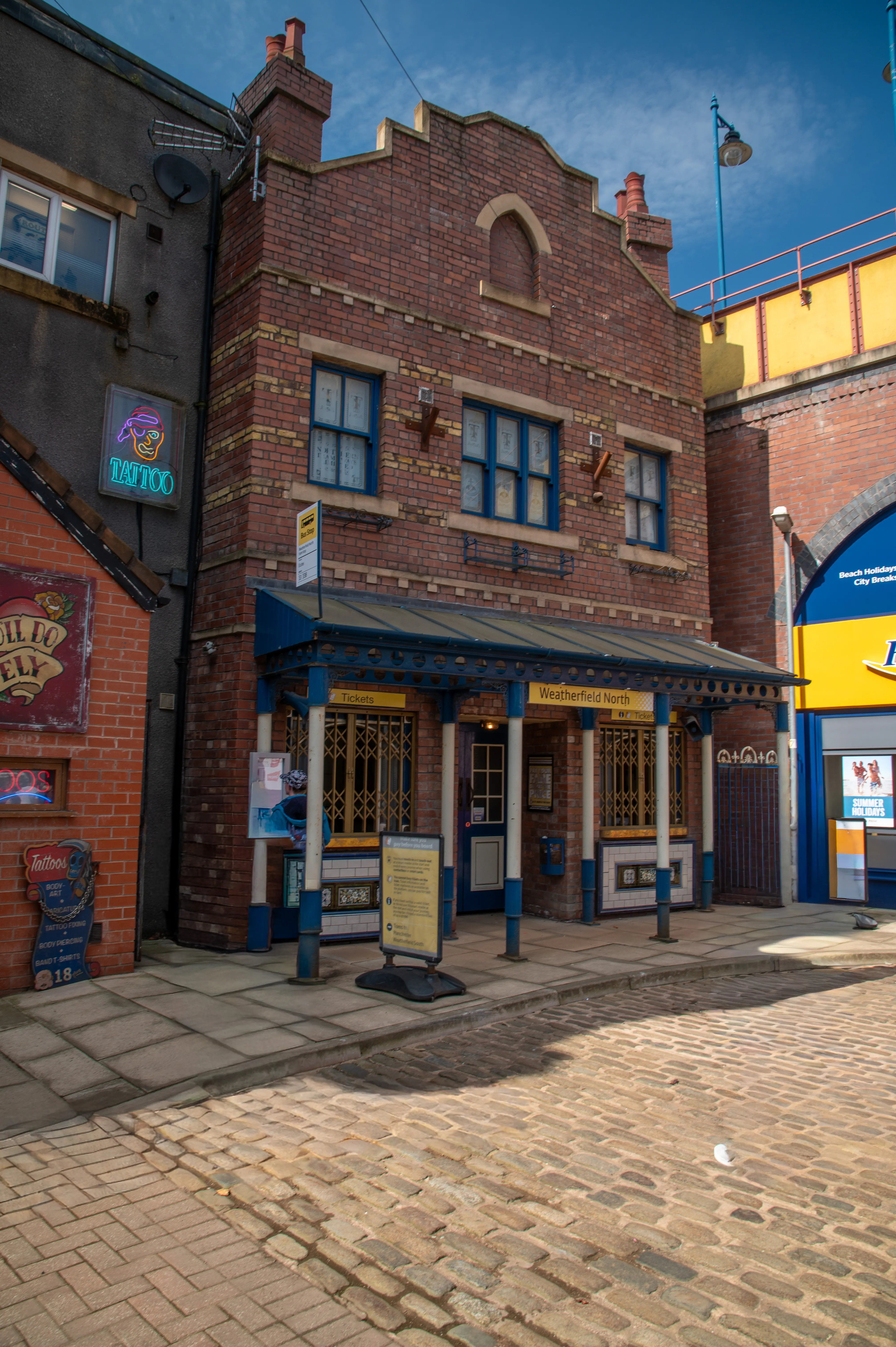 A brick building with a blue awning and &laquo;Weatherfield North Tickets&raquo; signage, set on a cobbled street. Two people stand in the foreground looking towards the building.