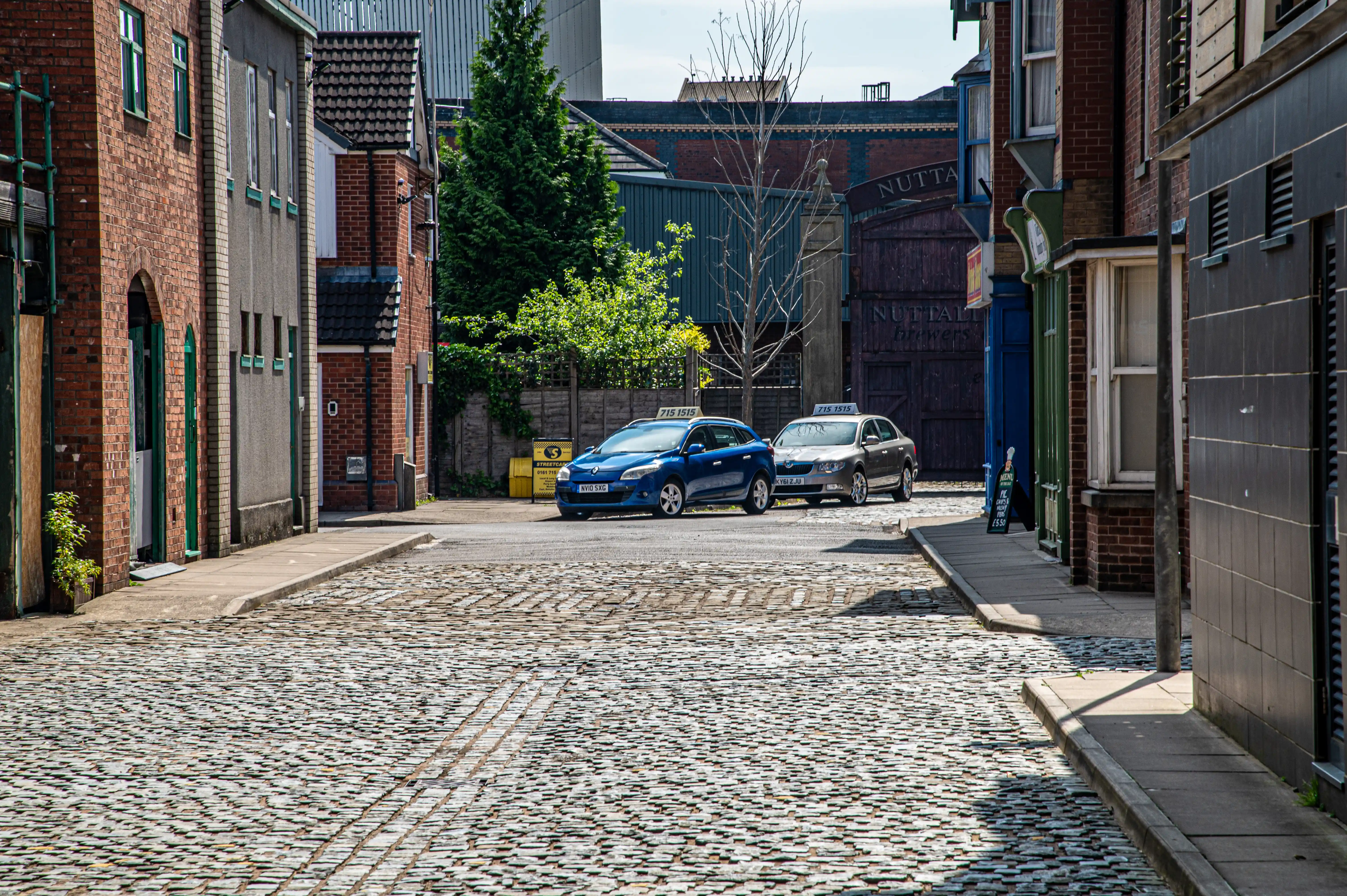 Two taxis, one blue and one silver, are parked on a street paved with cobbles. The street is flanked by brick buildings, some with green window frames and others with darker, modern facades. In the background, there are trees and industrial-style buildings