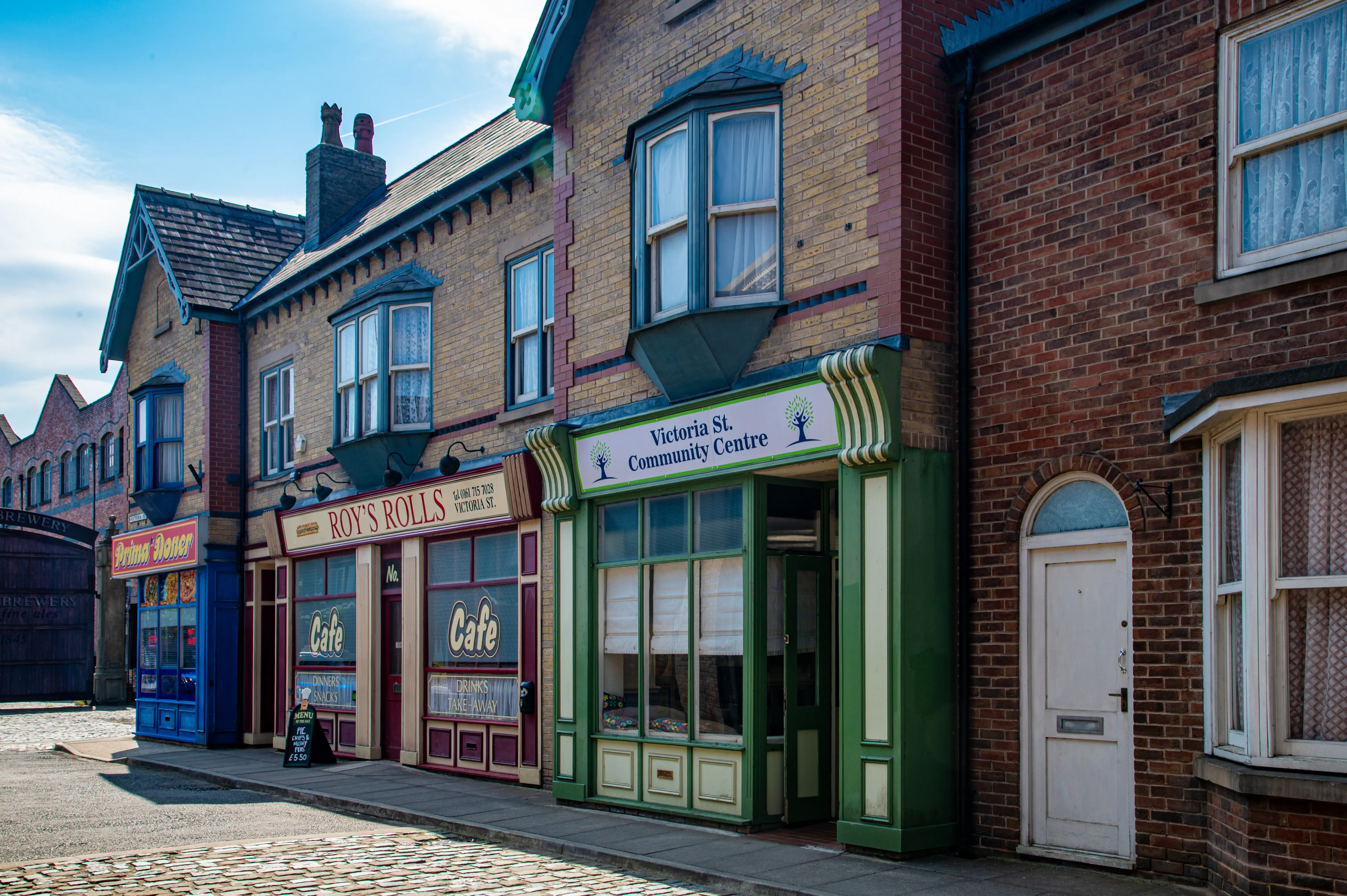 A picturesque street scene featuring the facade of a British television set, with brick buildings housing shops and a community centre, under a bright blue sky.