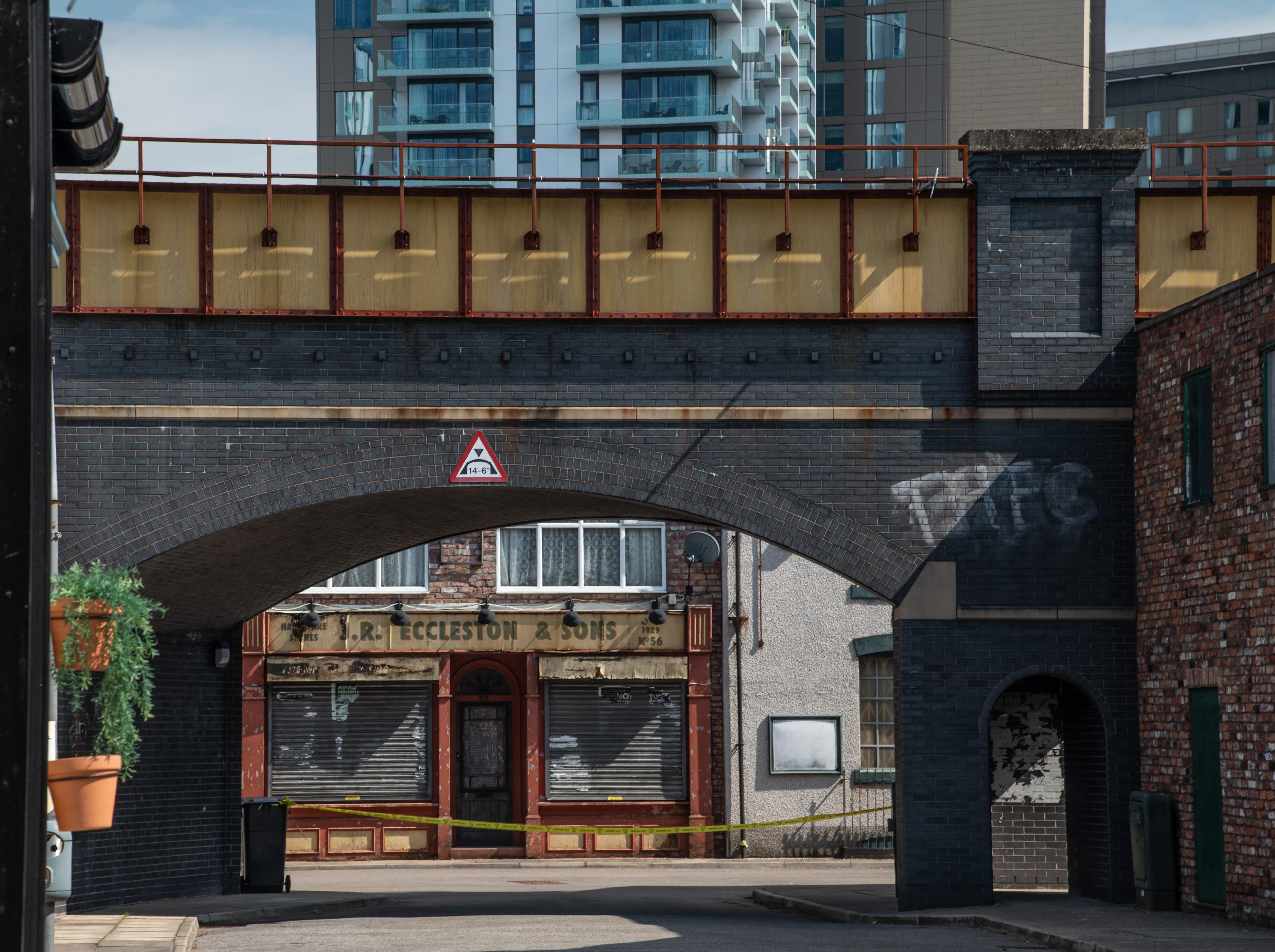 A Victorian brick railway viaduct arches over a street. Beneath the arch, a shop named 'J.R. Eccleston & Sons' has its shutters closed and is cordoned off with yellow tape. A modern building is visible in the background.