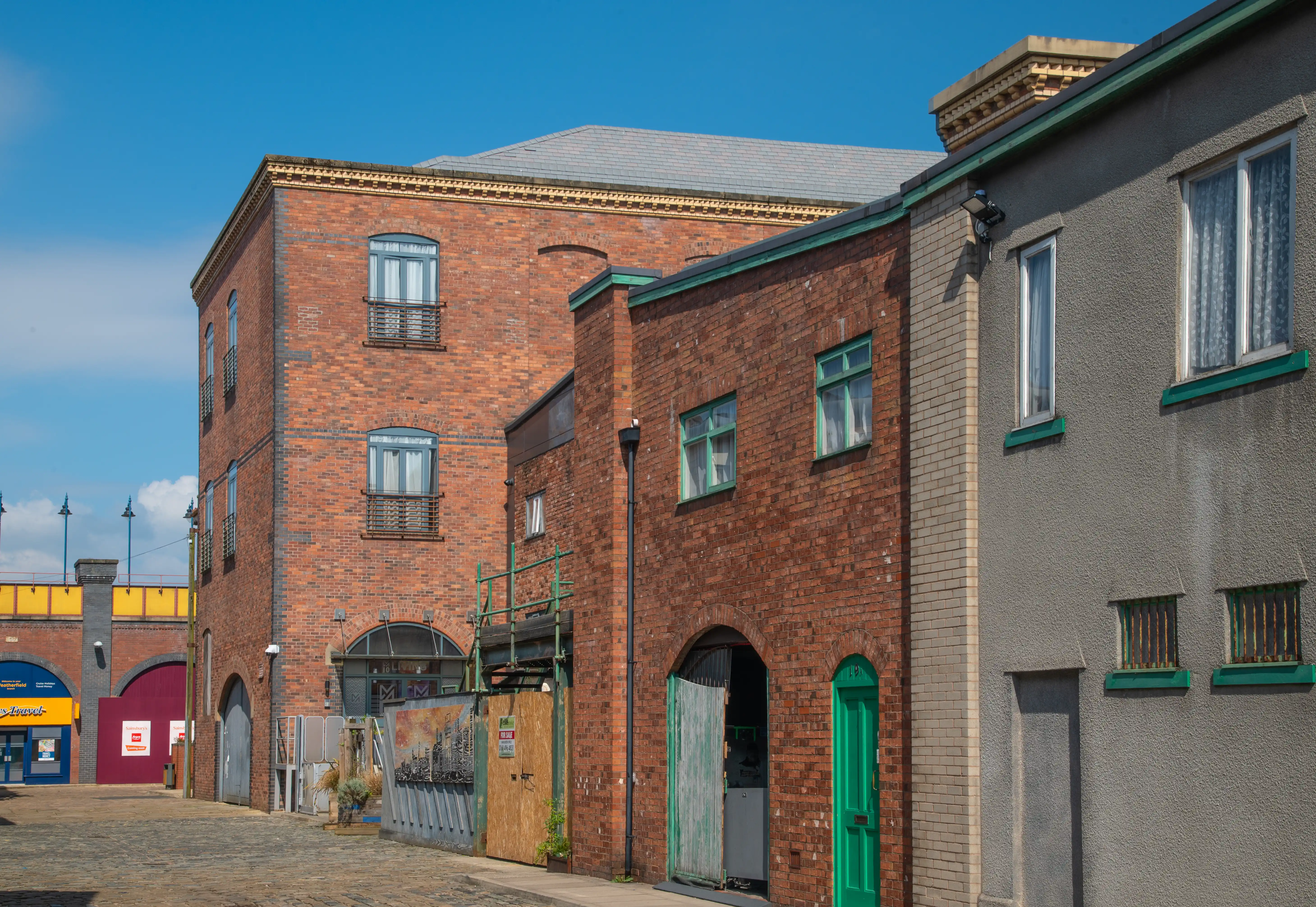 Red-brick industrial building next to a grey building with green window frames and a green door, viewed on a street under a clear blue sky. 