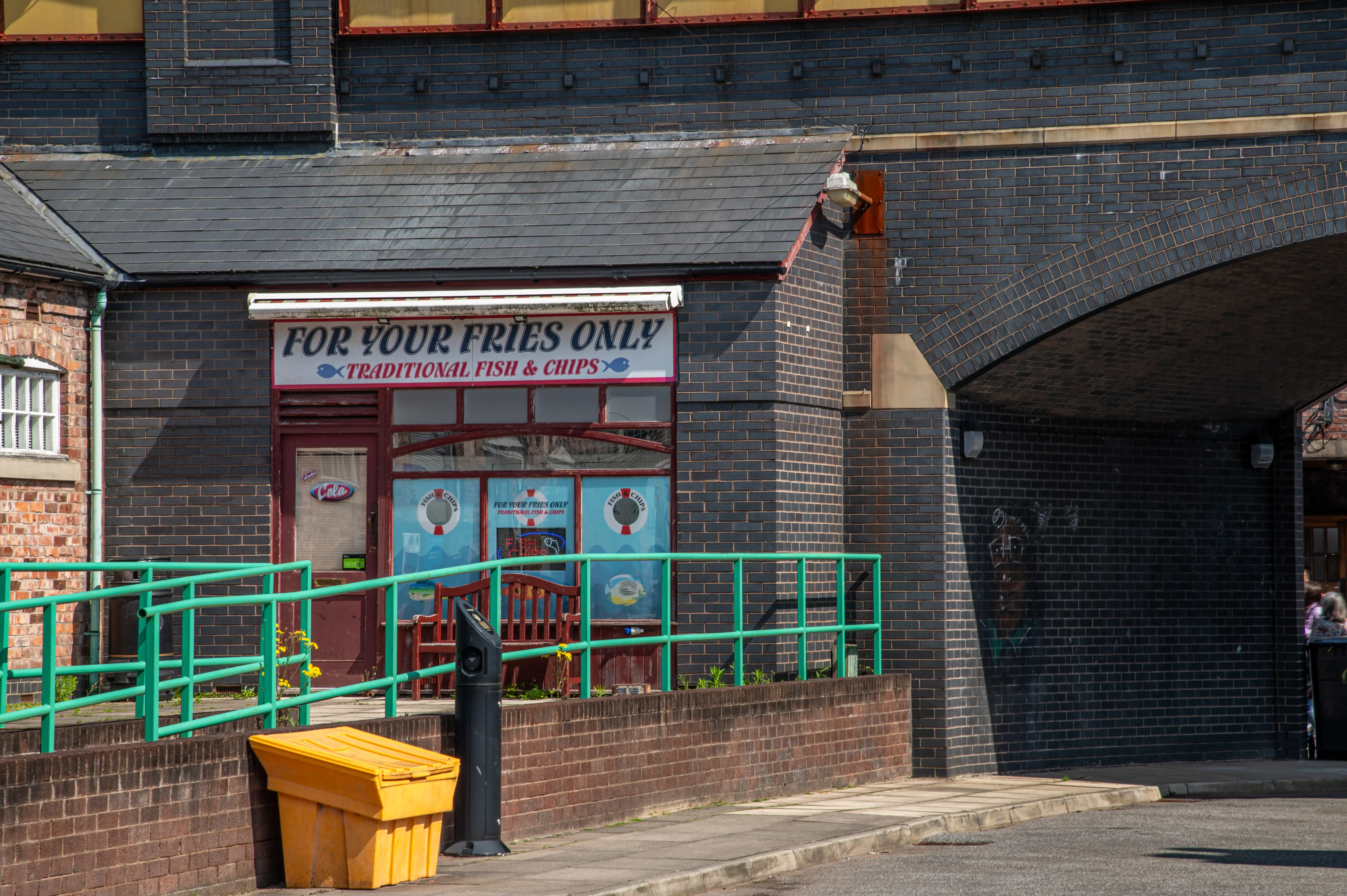 A quaint fish and chip shop named 'For Your Fries Only' is nestled beneath dark brick railway arches. Green railings and a yellow grit bin are visible.