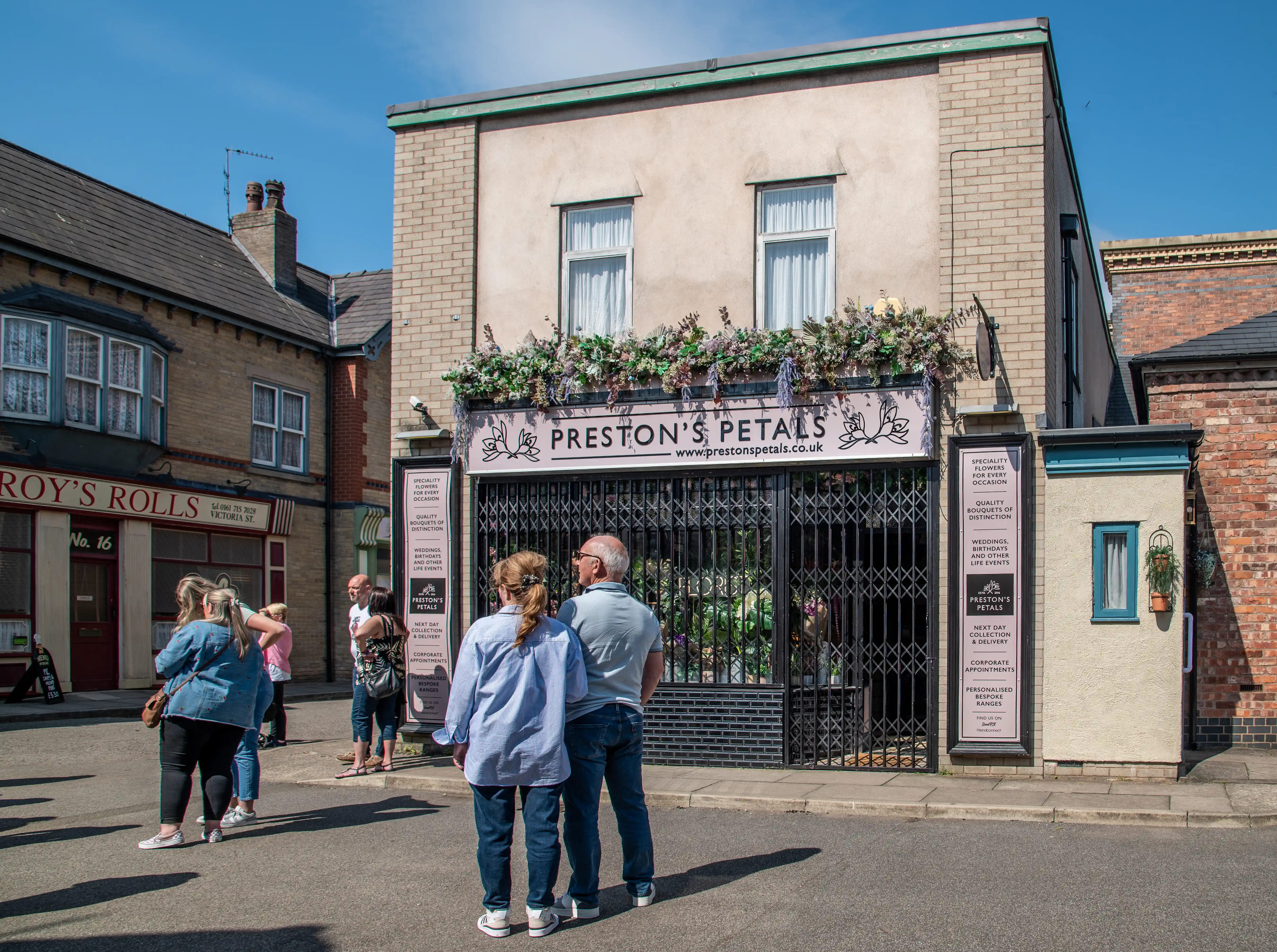 Two couples stand on a street, looking at the front of a flower shop called Preston's Petals. The shop has a large metal gate and floral decorations above the sign. Another shop, Roy's Rolls, is visible to the left