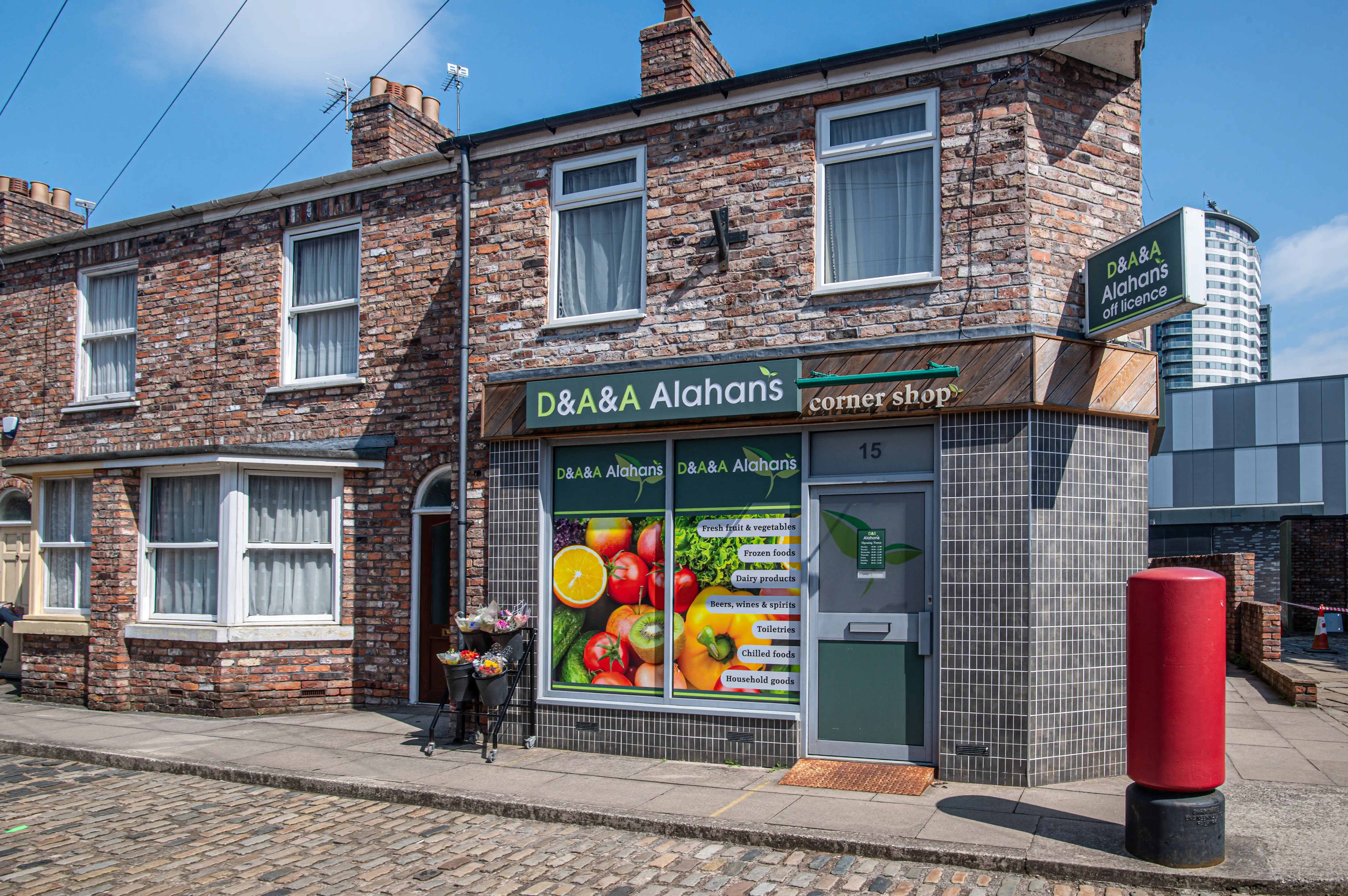 Exterior shot of D&A Alahan's corner shop from Coronation Street. The shop has a green sign with 'D&A Alahan's' and 'corner shop' printed on it. The shop window displays a variety of colourful fruits and vegetables. A red post box is situated on the pavement outside. The street is paved with cobbles, and the buildings are constructed of red brick. A modern skyscraper is visible in the background.