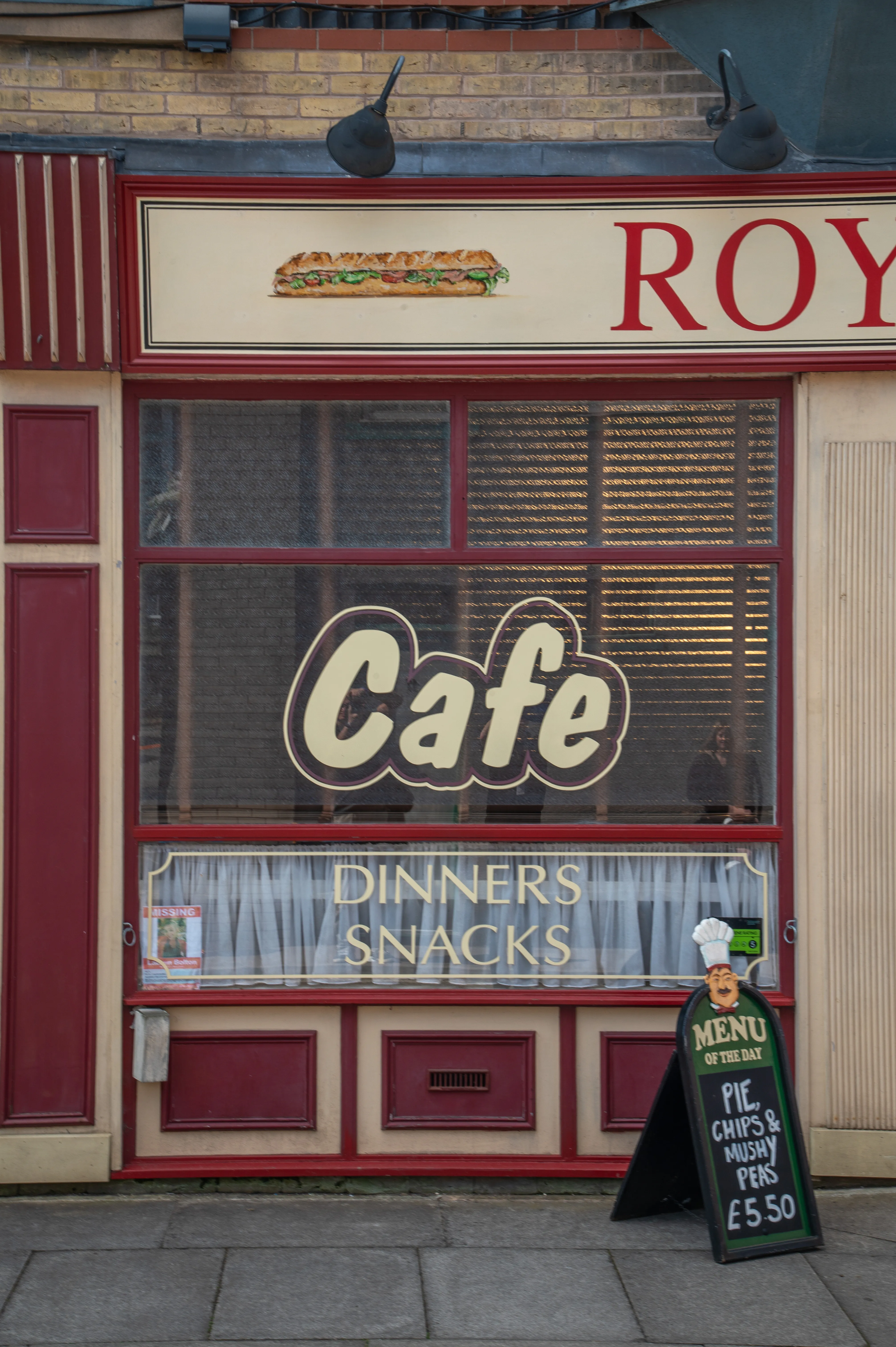 The front of a cafe with a large window displaying the word 'Cafe' and advertising 'Dinners & Snacks.' Above the window is a painted baguette. A sandwich board on the pavement advertises a pie, chips, and mushy peas meal deal.