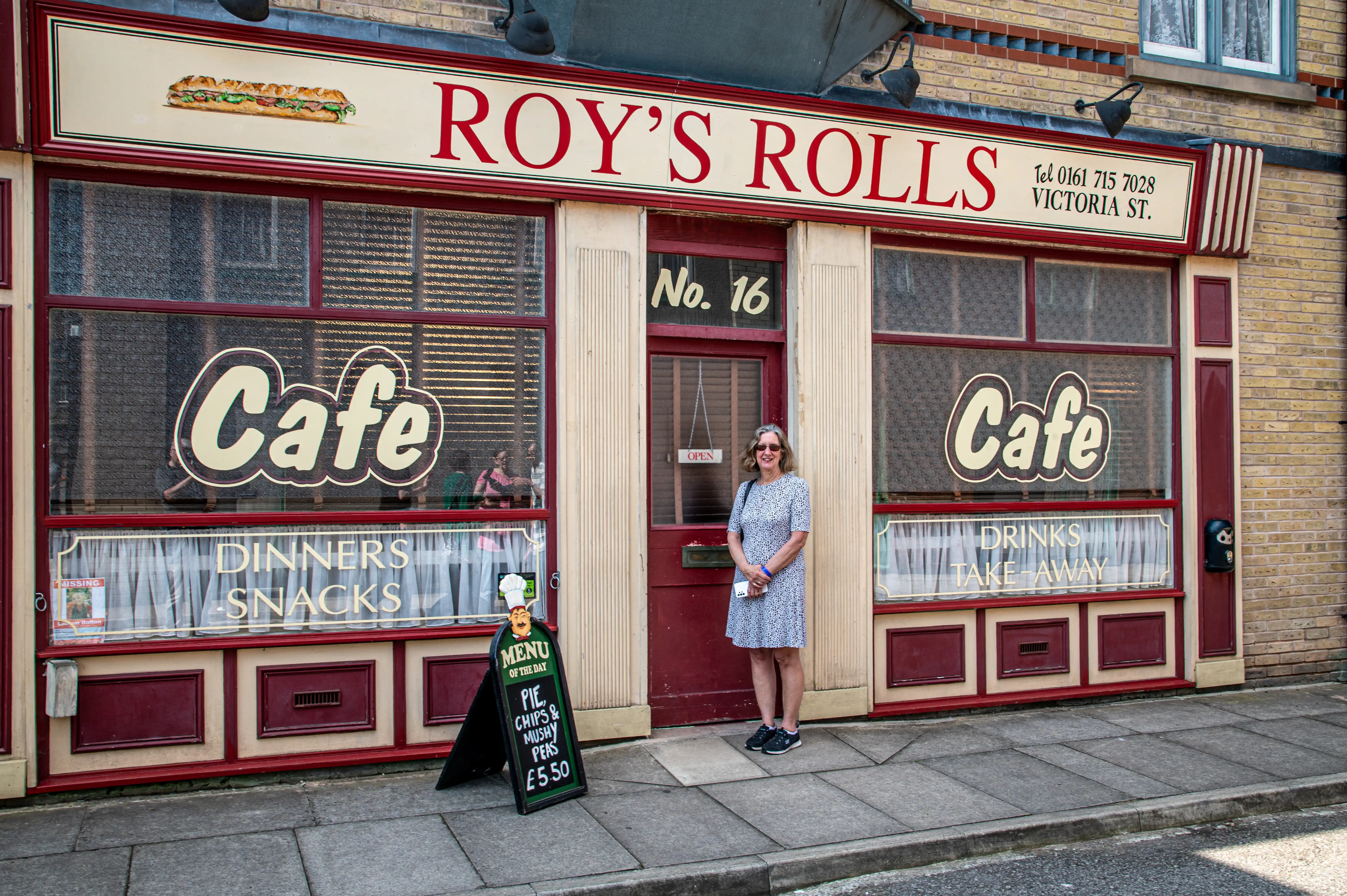 A woman in a blue floral dress stands on the pavement outside a cafe called &laquo;Roy's Rolls&raquo;. The cafe has large windows displaying signs for &laquo;Cafe&raquo;, &laquo;Dinners, Snacks&raquo;, and &laquo;Drinks, Take Away&raquo;. A blackboard menu is positioned in front of the cafe.