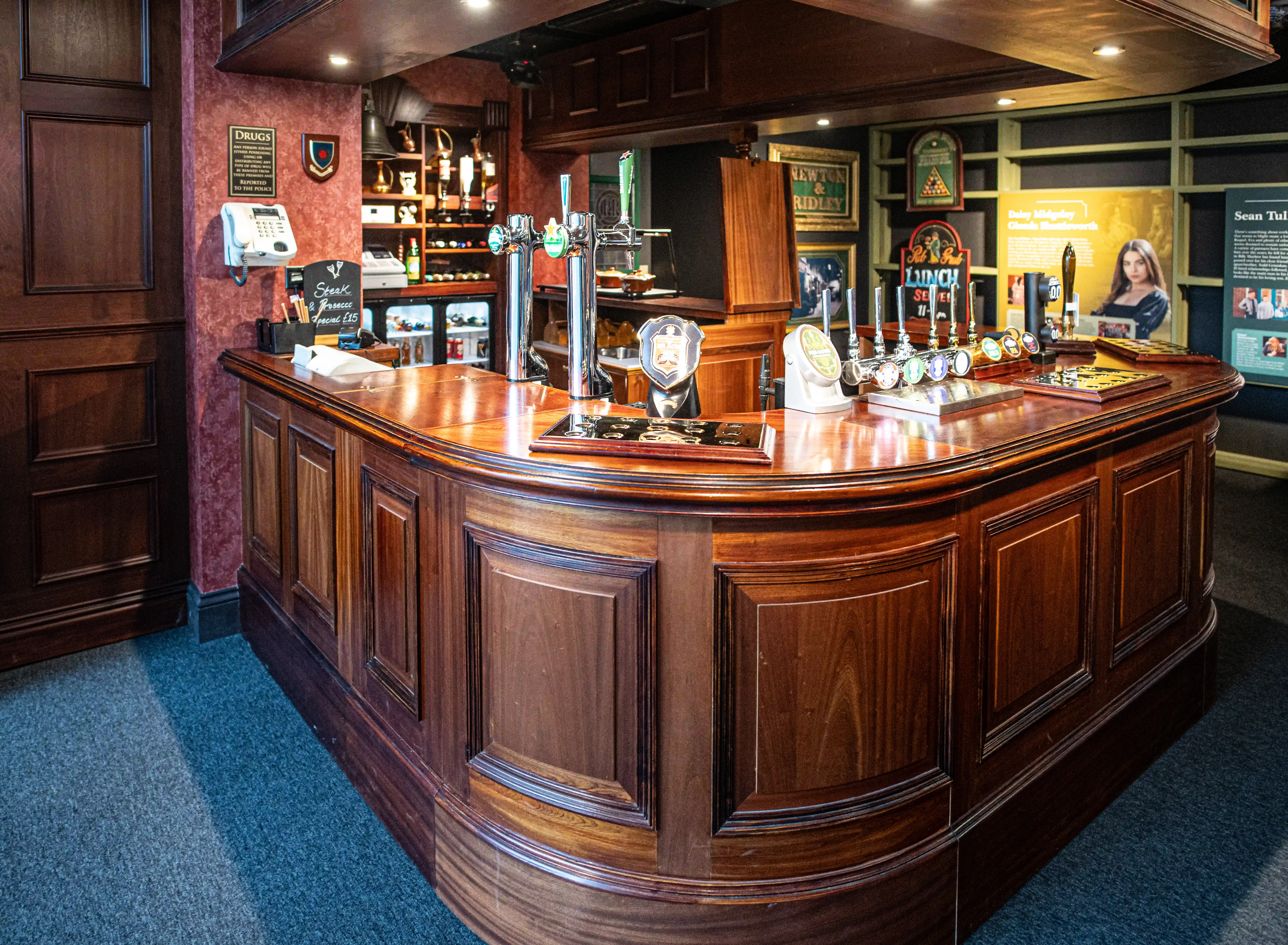 A polished dark wooden bar with ornate detailing, equipped with several brass beer taps. In the background, shelves display bottles and glassware, with framed pictures and informational panels on the wall, including a depiction of a woman.