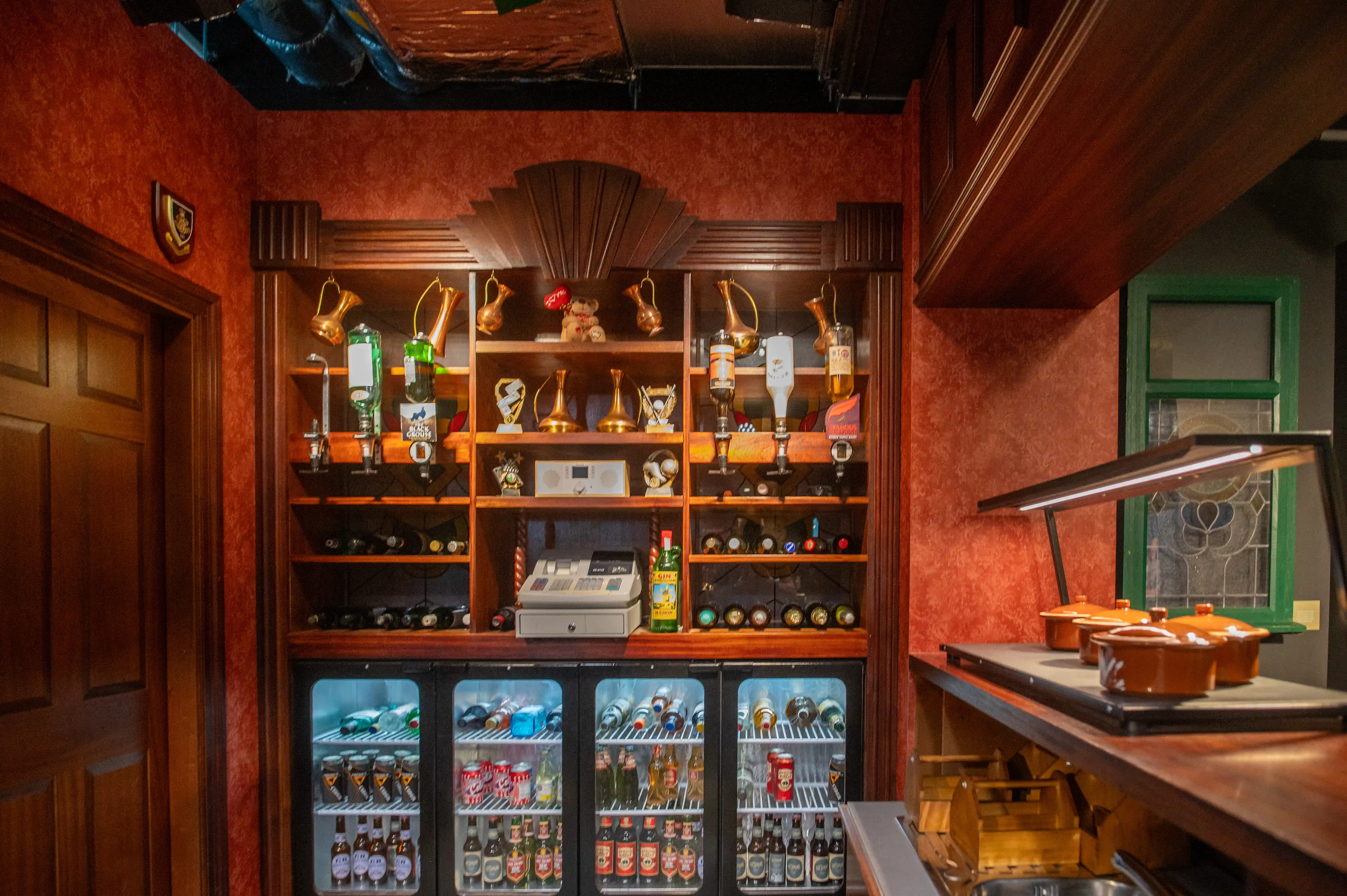 Interior view of a traditional pub bar, showcasing wooden shelving unit filled with alcoholic beverages, trophies, and drinks dispensers, with three under-counter refrigerators below, and a counter with warmers in the foreground.
