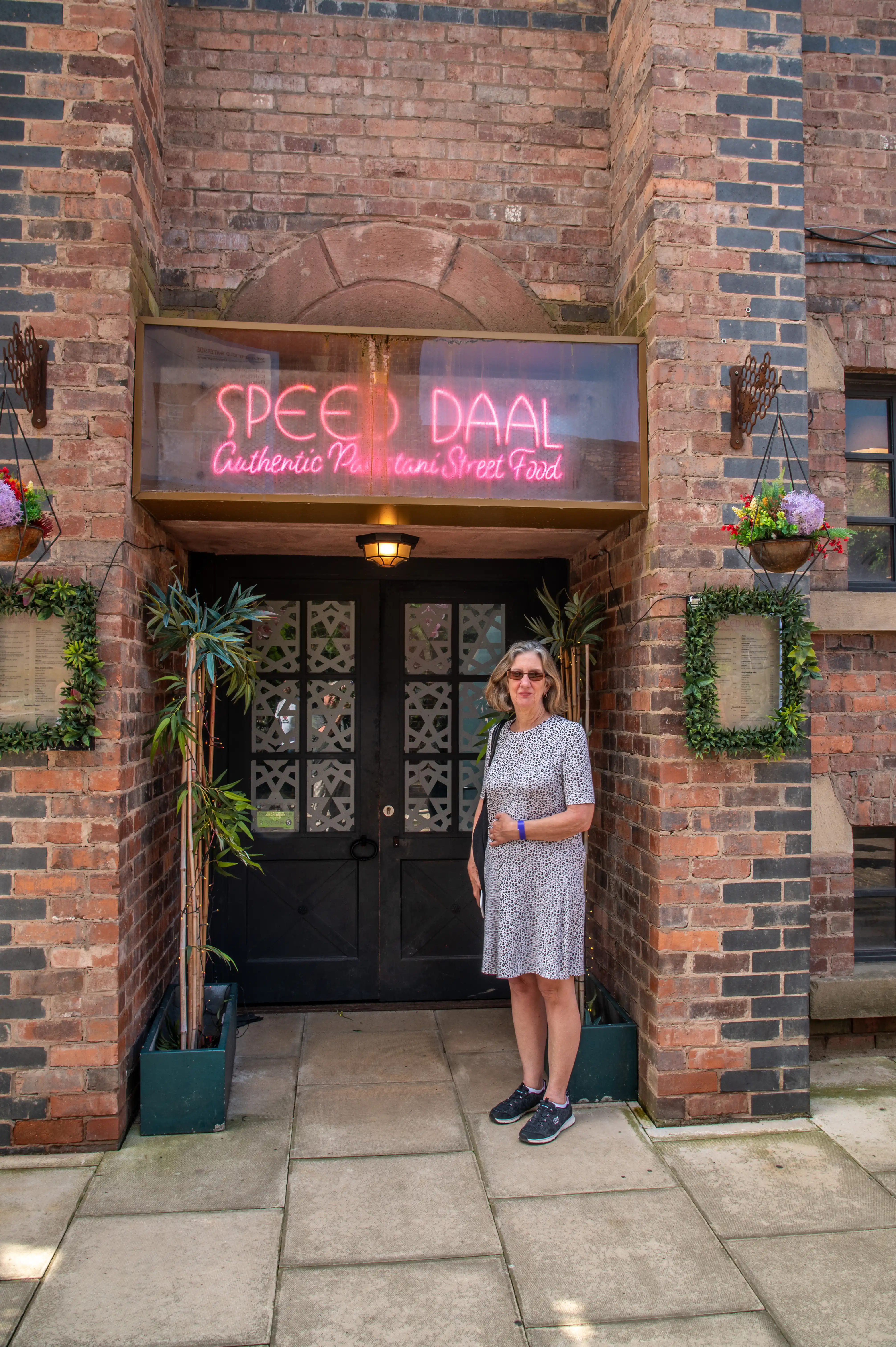 A woman in a patterned dress stands on a paved walkway in front of a brick building with a neon sign reading 'Speed Daal Authentic Pakistani Street Food'.