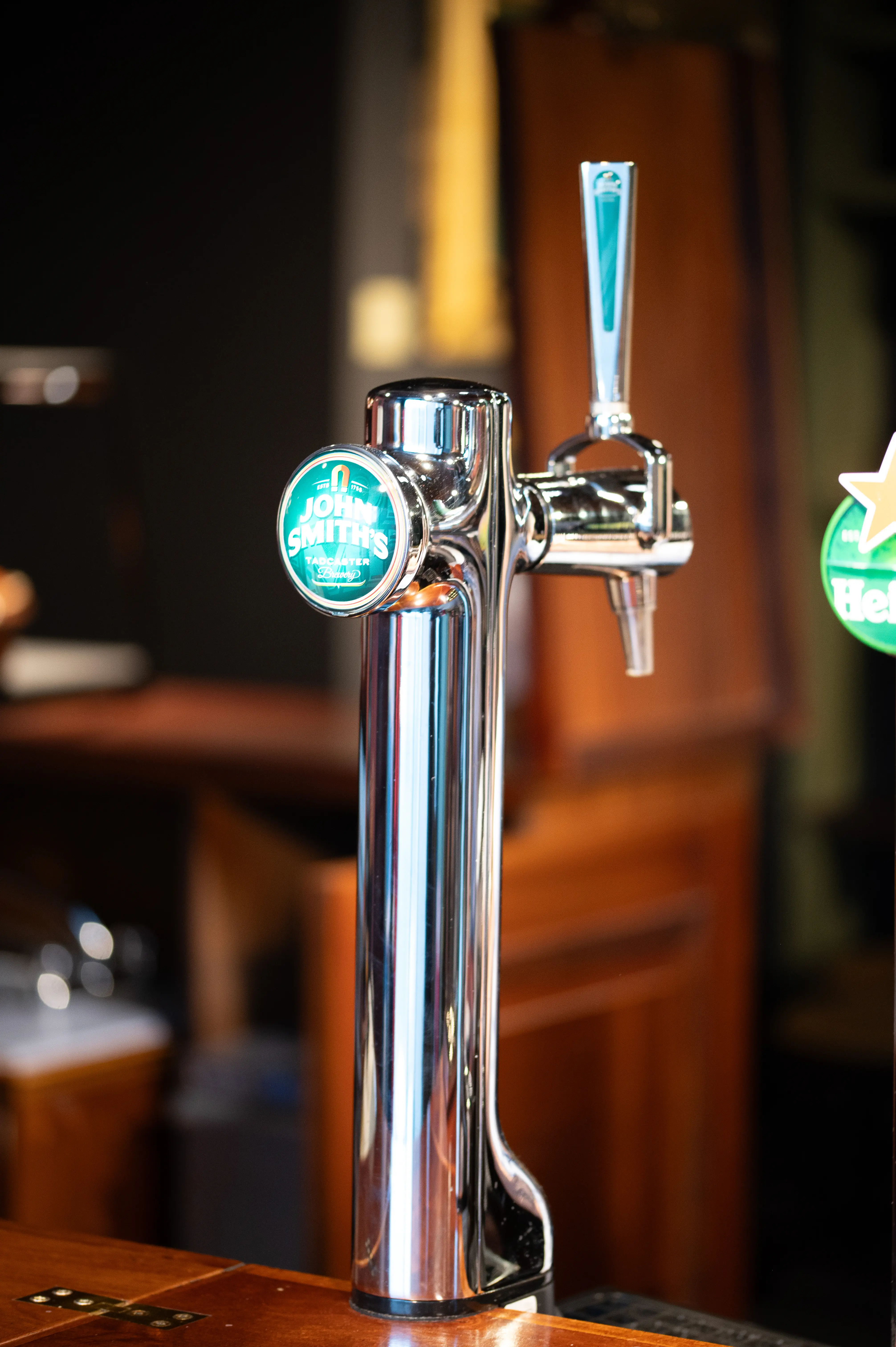 A gleaming chrome beer tap with a circular green 'John Smiths' logo is prominently displayed. The tap is positioned on a polished wooden bar, with a blurred background of a pub interior.