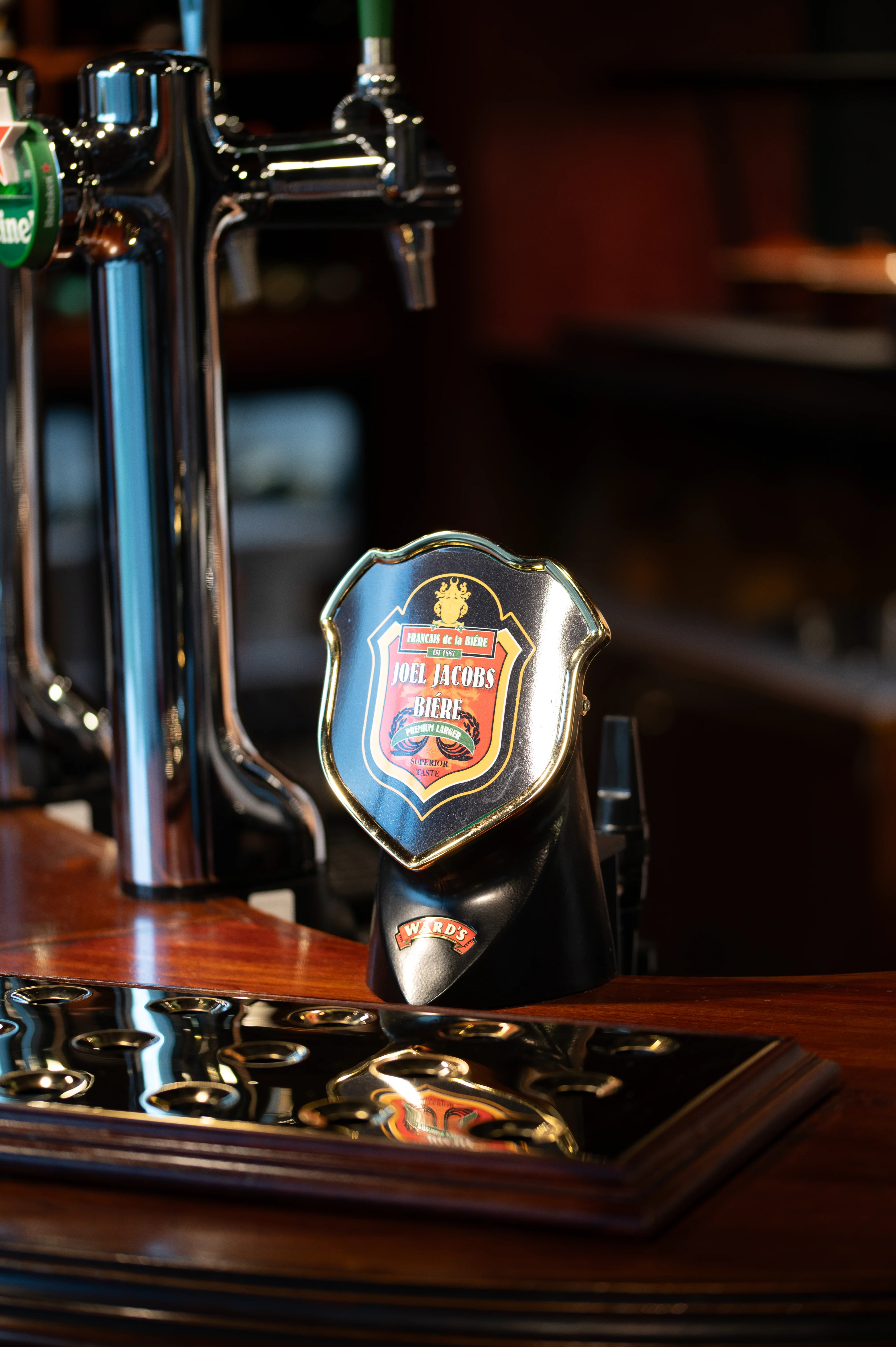  Close-up of a black beer jug with a crest label and gold trim on a wooden counter in a pub interior.  