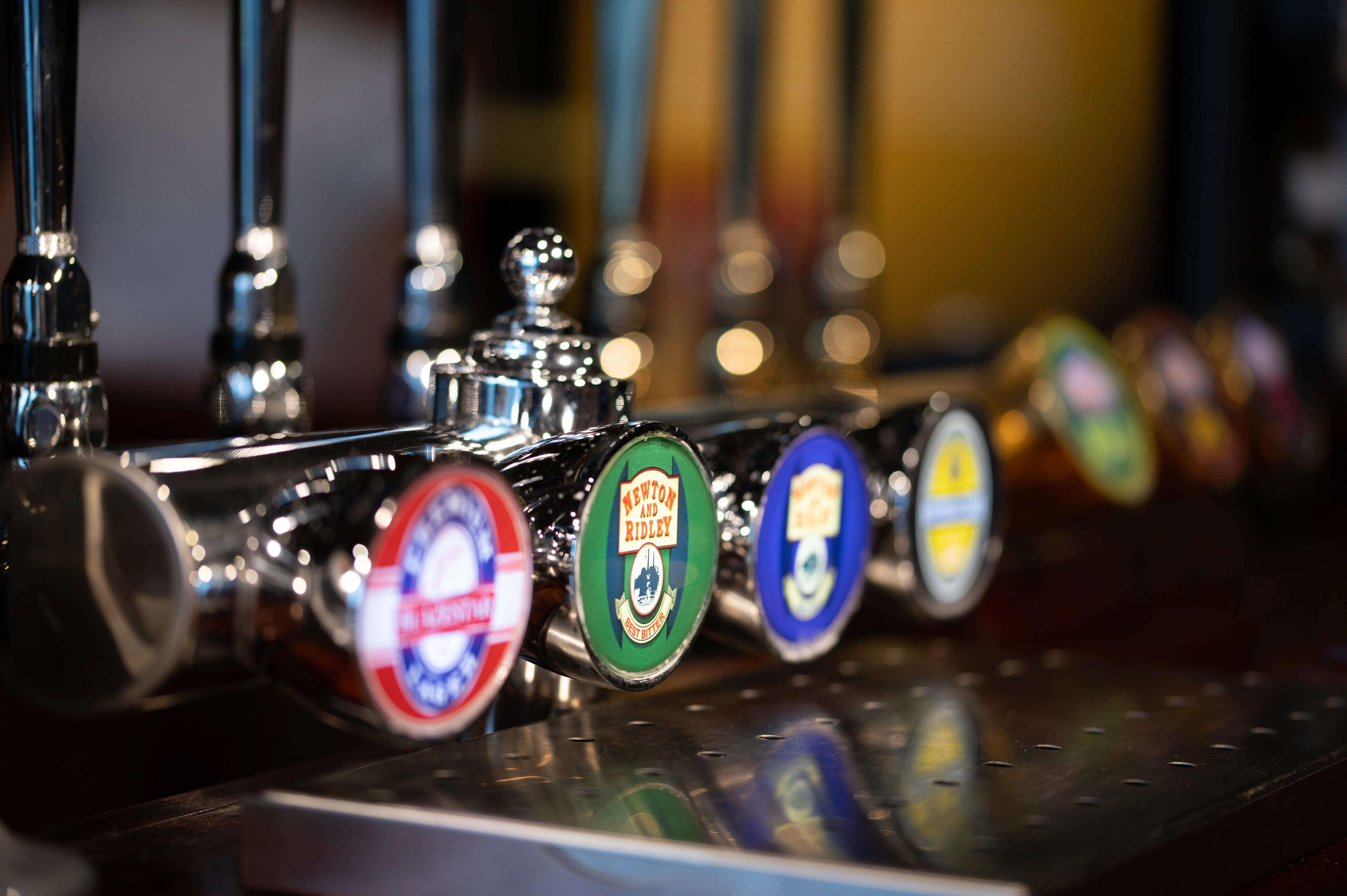 A row of shiny chrome beer taps in a pub, each with a circular pump clip featuring different logos and colours. The tap on the far left has a red and white clip, followed by a green clip with 