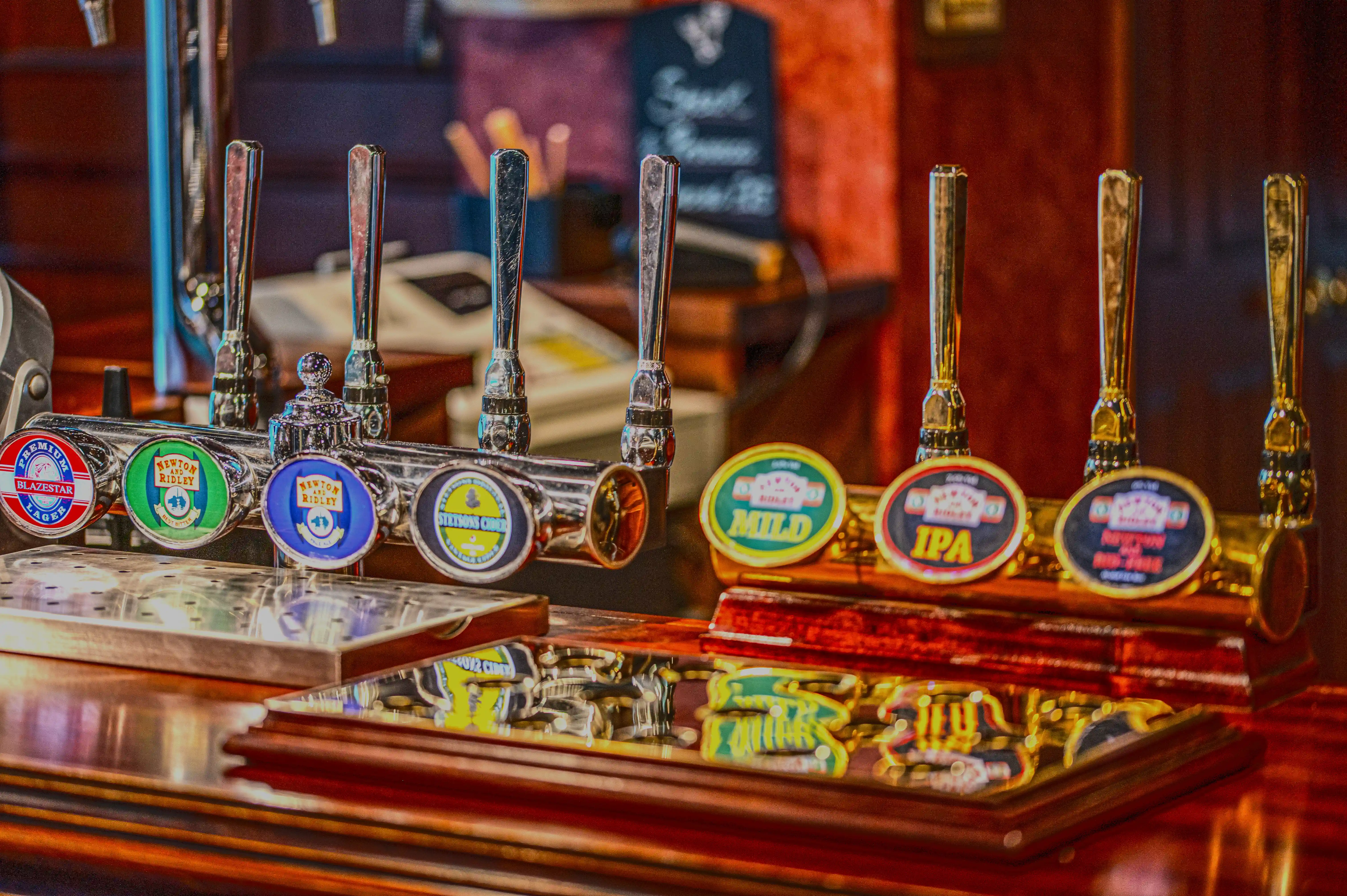 A close-up shot of several polished chrome beer taps with decorative pump clips displaying different ale names such as 'Mild' and 'IPA' on a wooden bar in a pub.