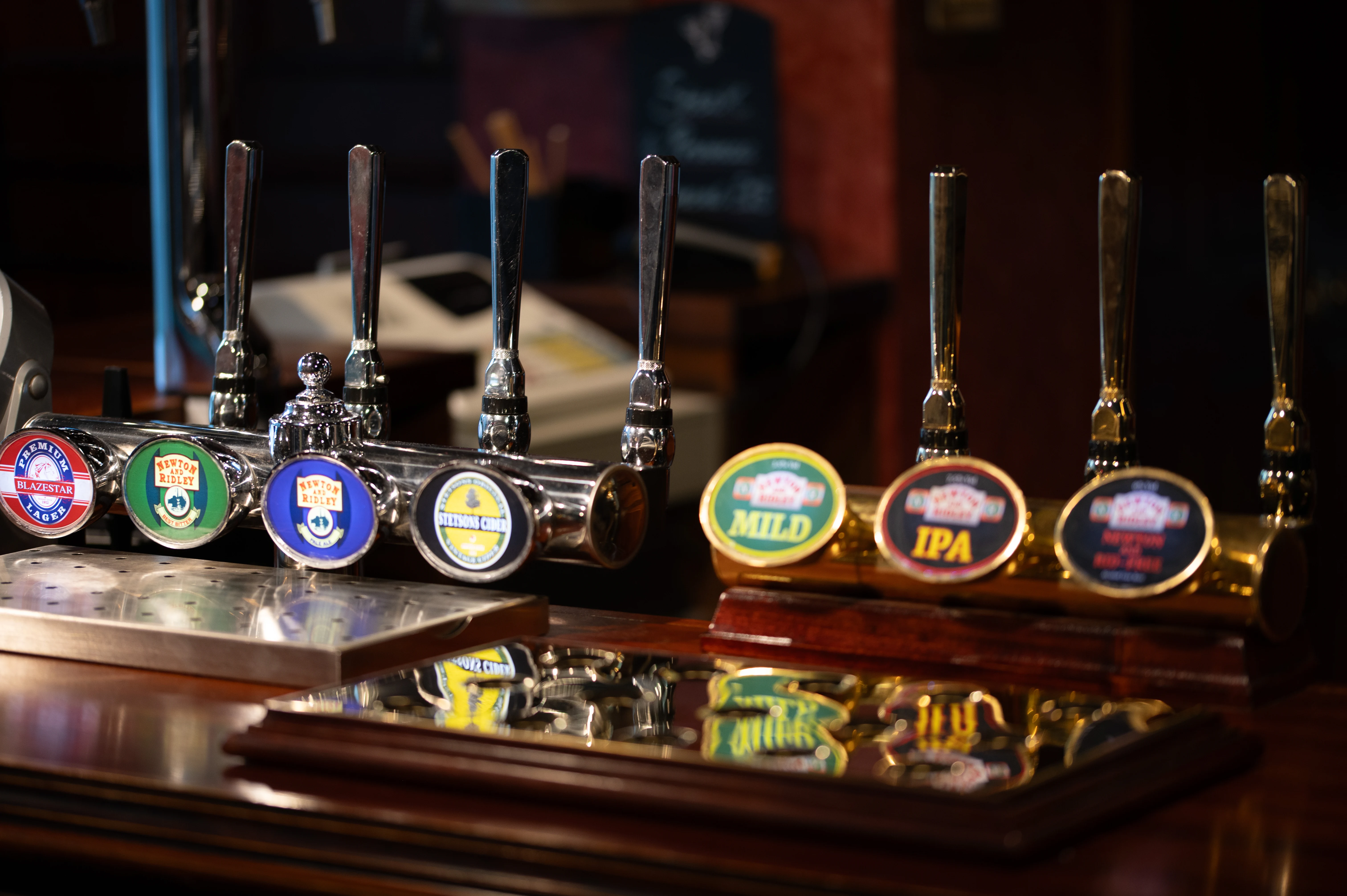 A close-up shot of several polished chrome beer taps with decorative pump clips displaying different ale names such as 'Mild' and 'IPA' on a wooden bar in a pub.