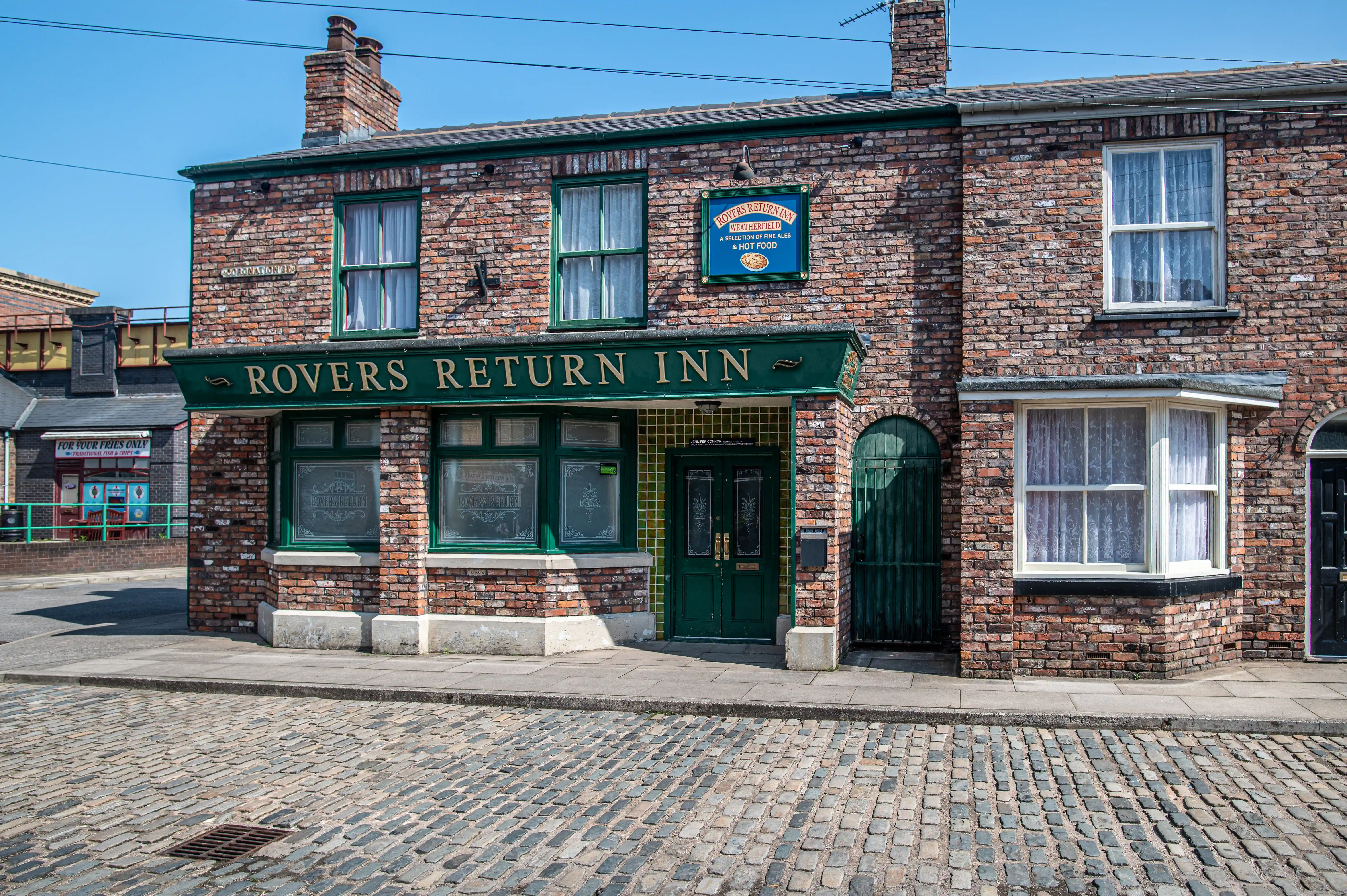 Exterior of the Rovers Return Inn, a brick building with a green fascia, double green doors, and bay windows, situated on a cobblestone street.