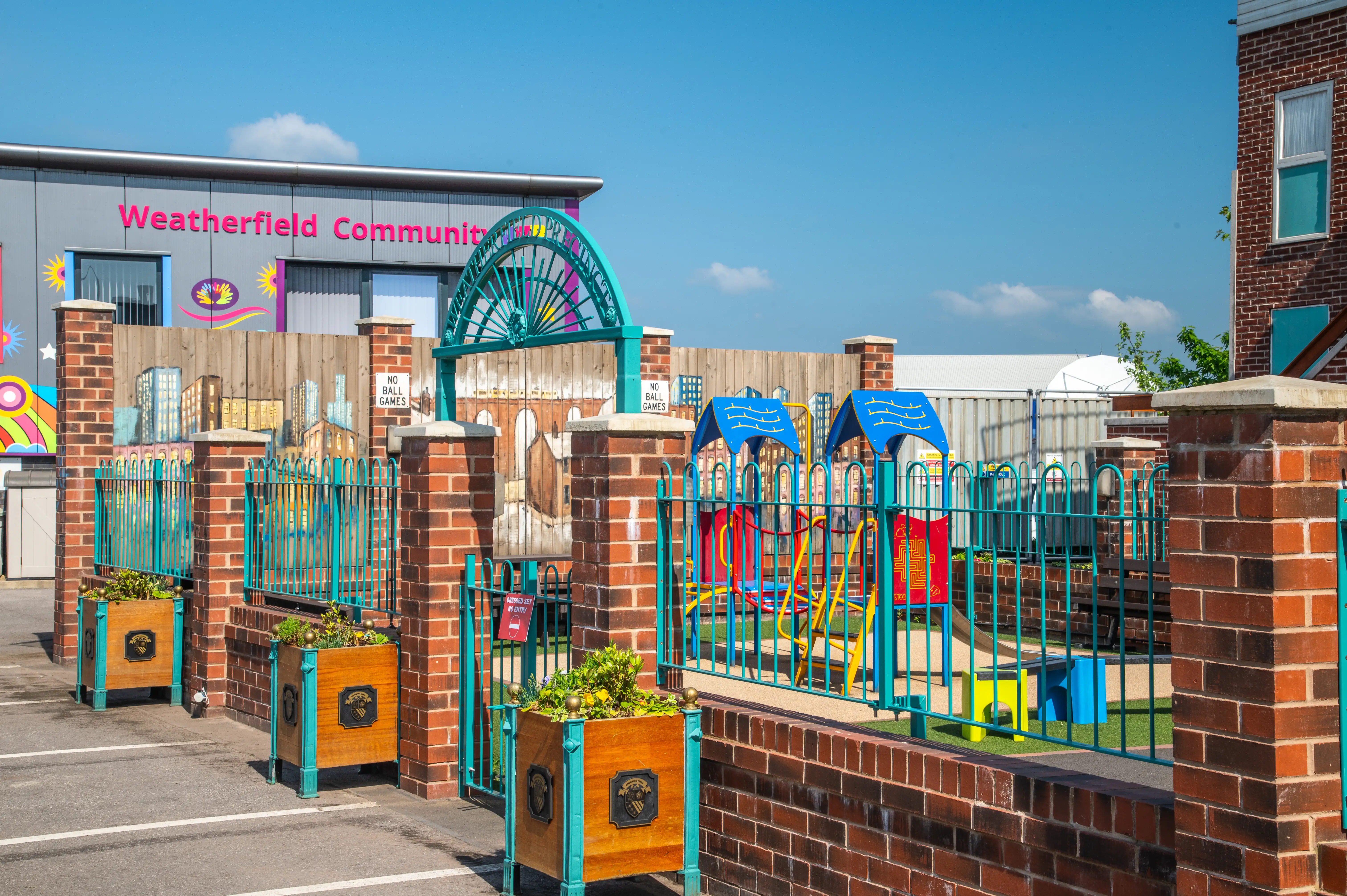 A children's playground with blue-roofed play structures and a teal safety fence, set against the backdrop of a modern building labelled 