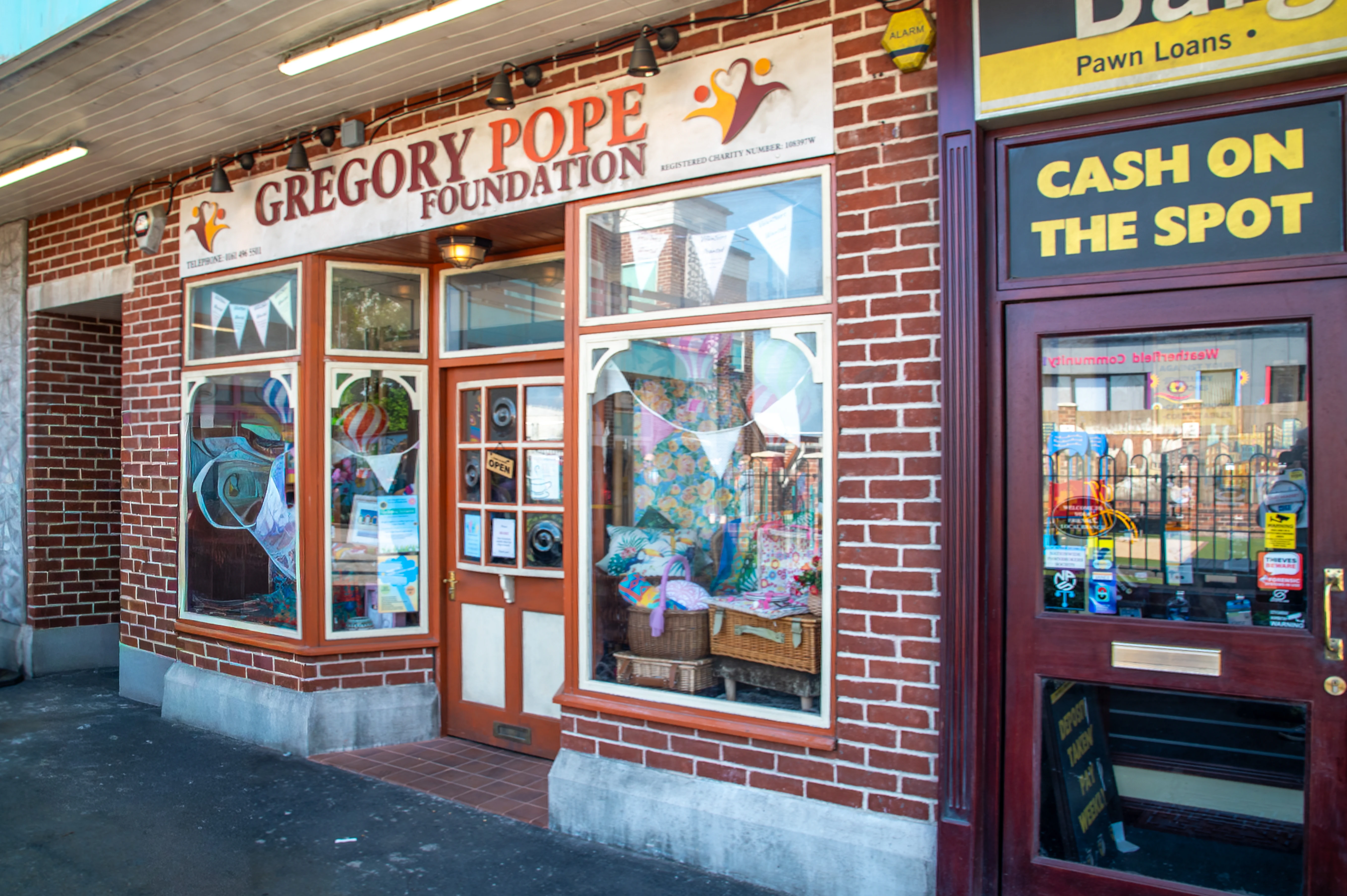 The exterior of the Gregory Pope Foundation shop, featuring a brick facade, large display windows showcasing various items, and a prominent sign. Adjacent to it is a pawn shop.