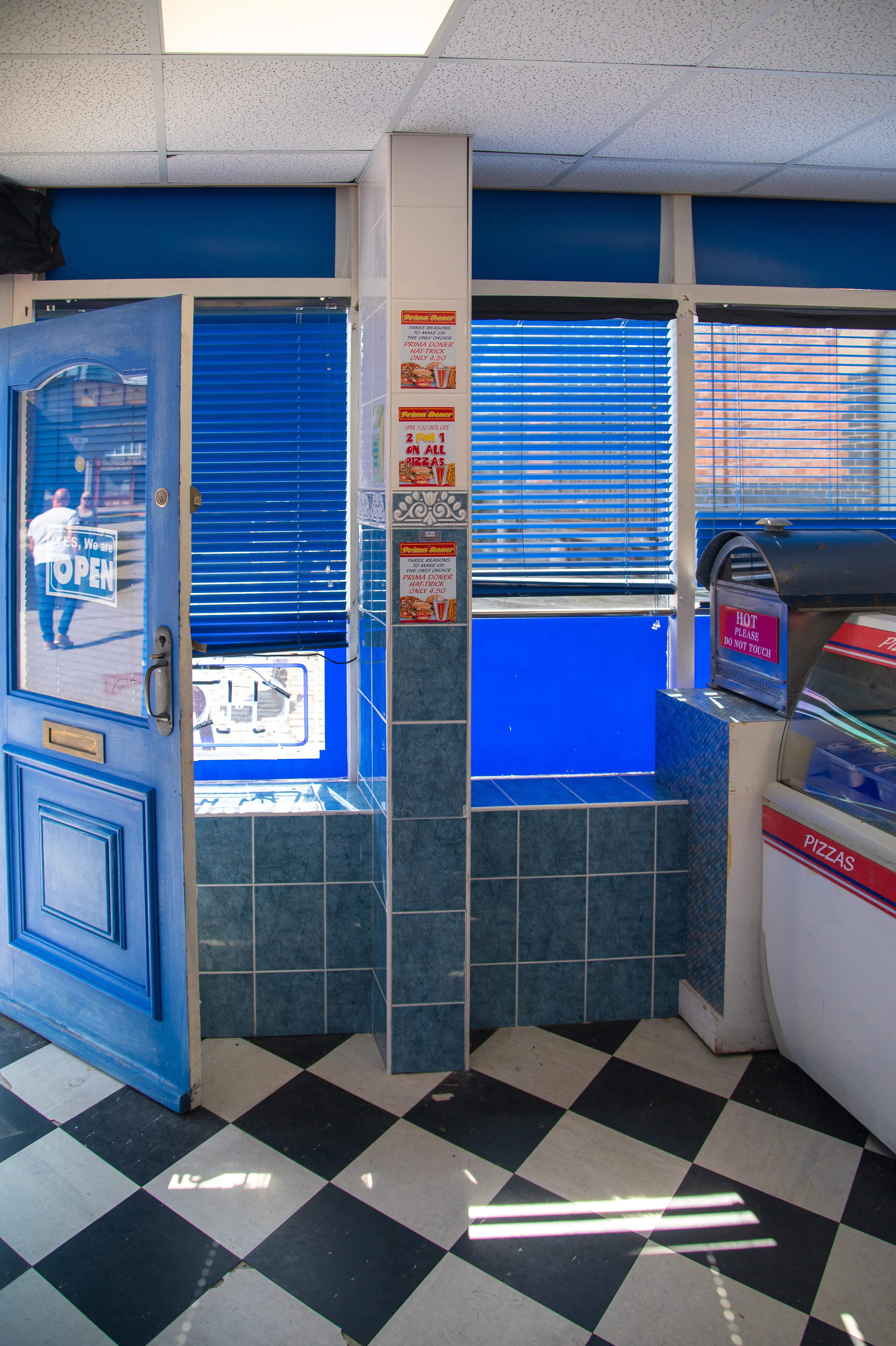 Interior view of a takeaway shop with a blue door, blue blinds, blue tiled walls, and a black and white chequered floor.