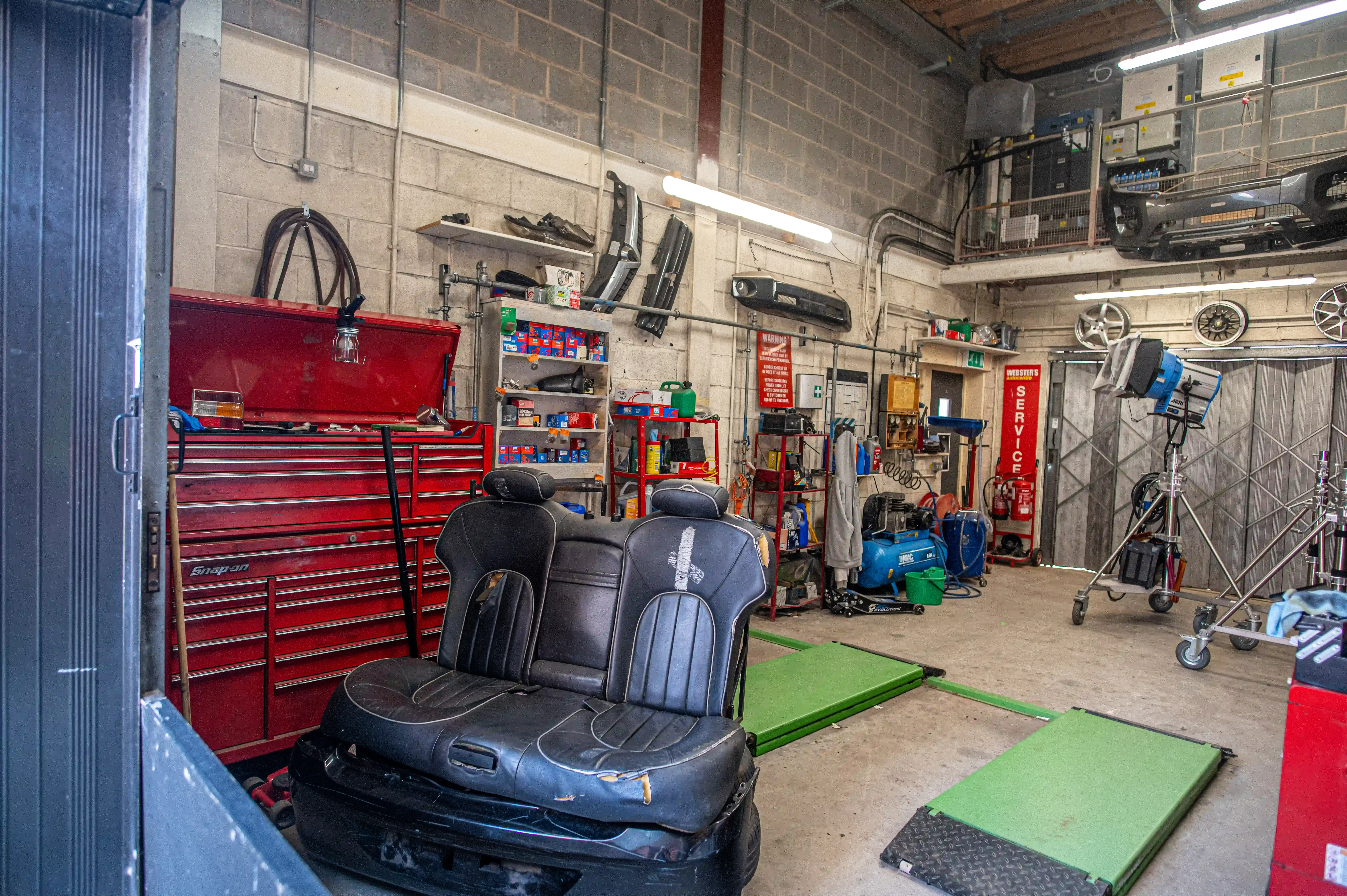 Workshop interior with red tool cabinet drawers, shelves of tools, and two black vehicle seats on a green mat.  