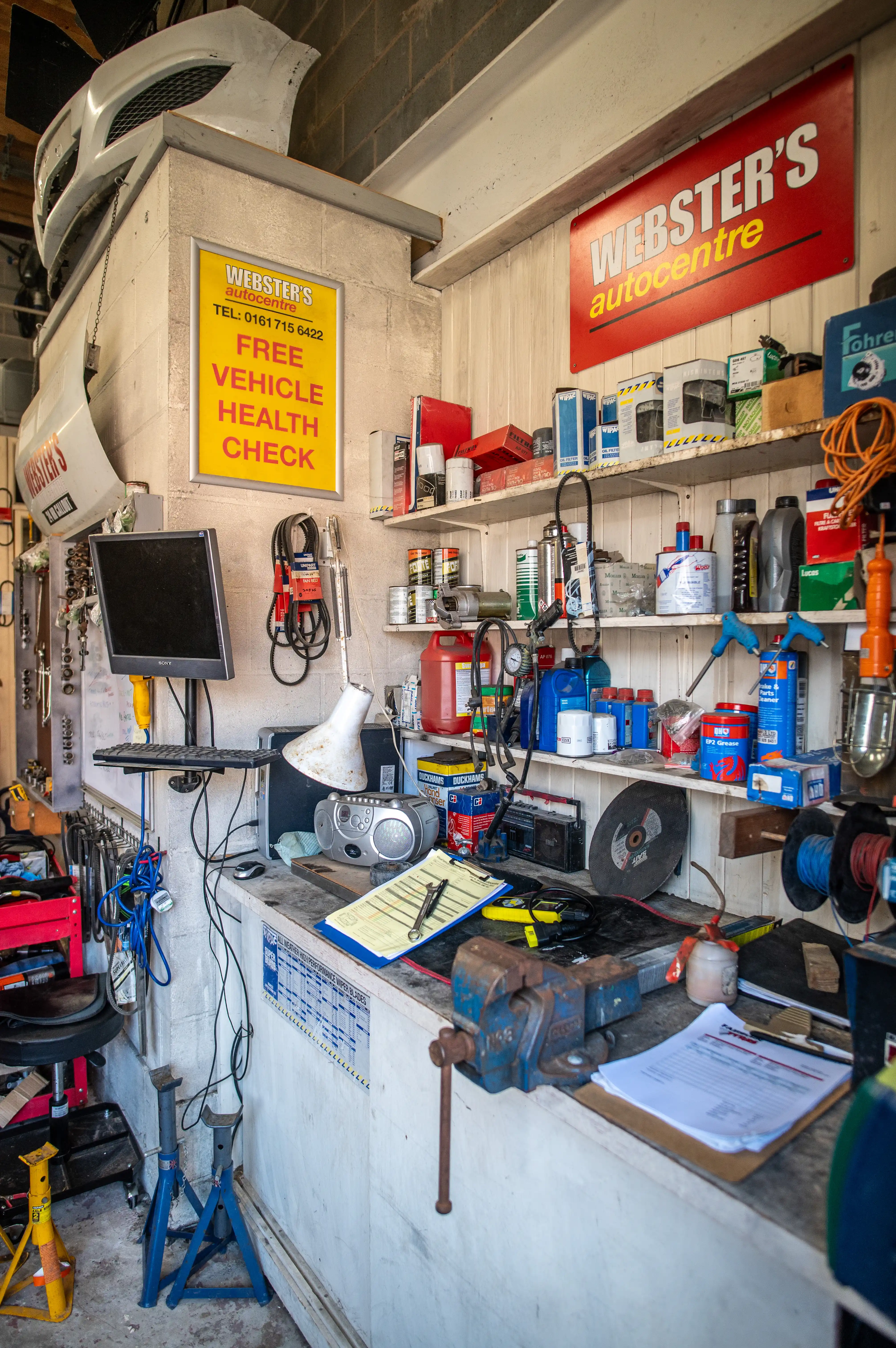 Interior view of a cluttered vehicle repair workshop, showing a workbench covered in tools, fluids, and paperwork, with shelves stocked with automotive supplies and a prominent sign advertising a Free Vehicle Health Check.
