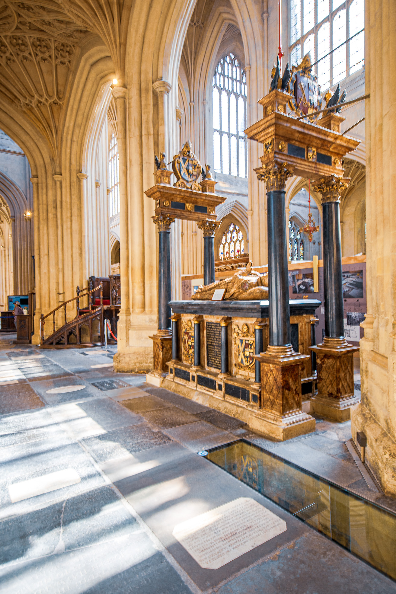 The image depicts an ornate interior of a historic cathedral or church, featuring intricate Gothic architecture. The focal point is a large, elaborately decorated tomb or monument, likely of significant historical importance. The monument is surrounded by tall, intricately carved pillars and arches, with detailed stonework and stained glass windows in the background. The floor is made of polished stone, and there are informational plaques placed in front of the monument. The overall atmosphere is one of reverence and historical significance.