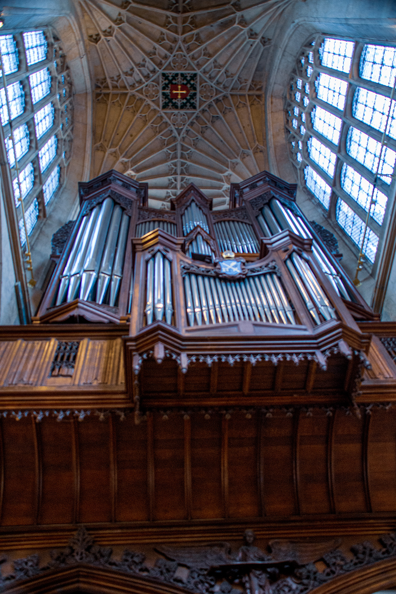 The image depicts the interior of a grand cathedral, focusing on an ornate pipe organ situated high above the wooden paneling. The ceiling features intricate stonework and large stained-glass windows that allow natural light to illuminate the space, creating a serene and majestic atmosphere.